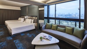 Habitación de hotel con cama, sofá, mesa de centro y un amplio ventanal con vistas a la ciudad.