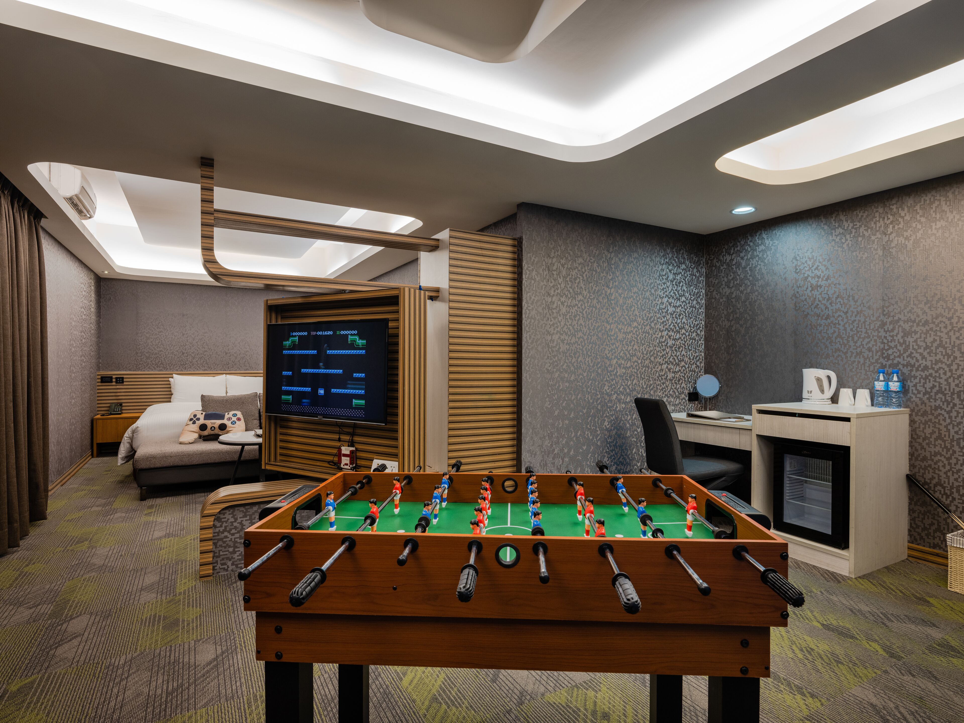 Habitación de hotel con mesa de fútbol, televisor, escritorio con computadora y cama.