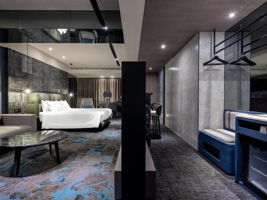A modern hotel room with a large bed, a seating area with a round table, a fireplace, and a wardrobe with a mirror.