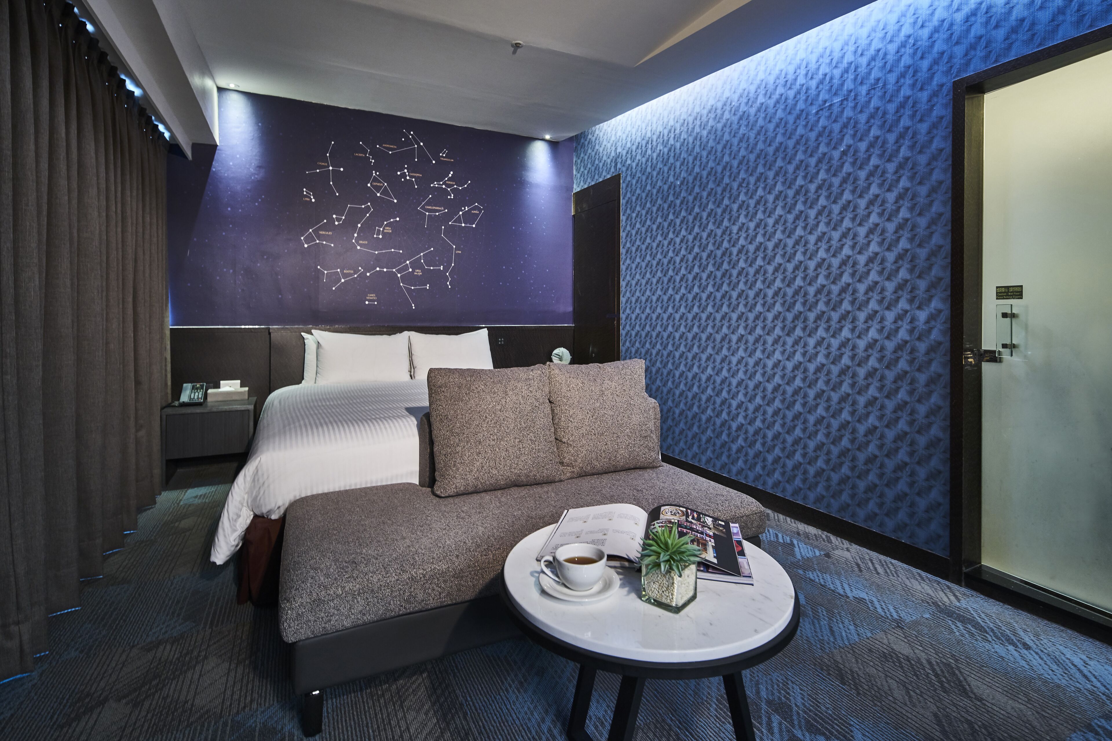 Una habitación de hotel moderna con una cama grande, un pequeño sofá, una mesa redonda con una taza y una planta, y una pared con un diseño geométrico.