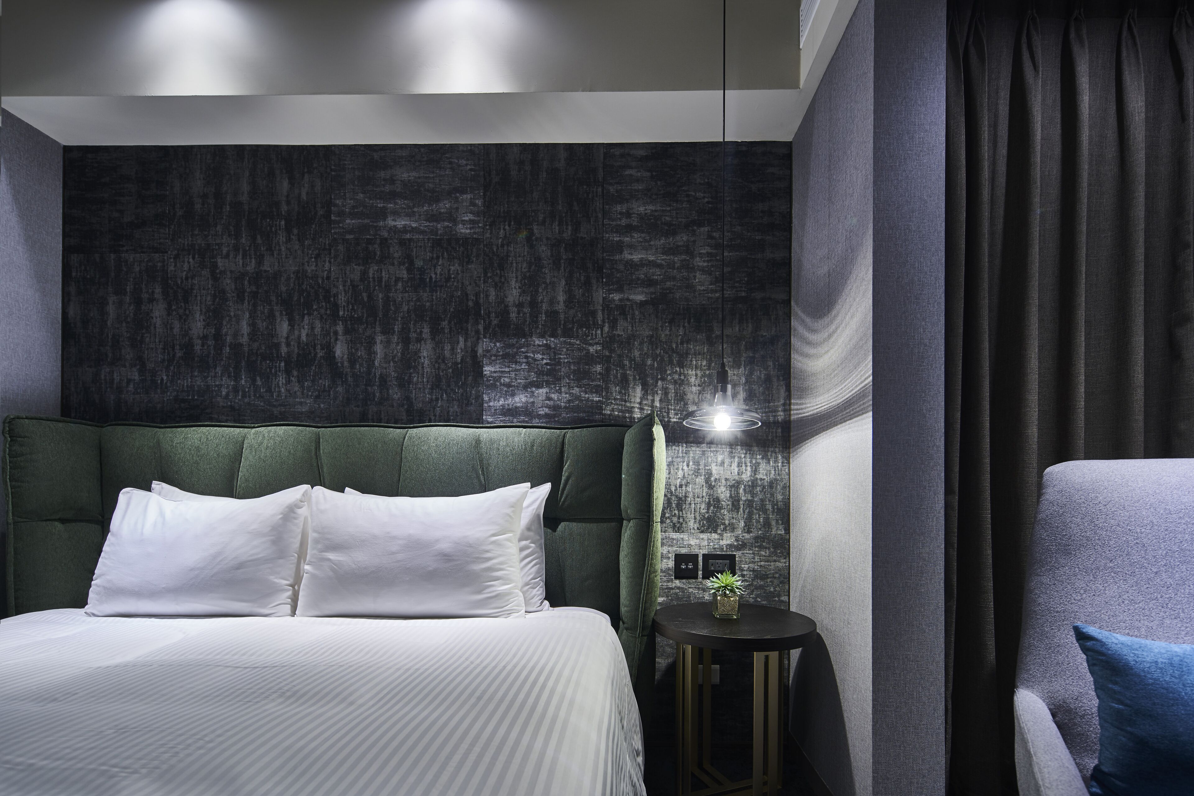 A modern hotel room with a large bed, a green upholstered headboard, a small round table with a plant, and a large window with curtains.