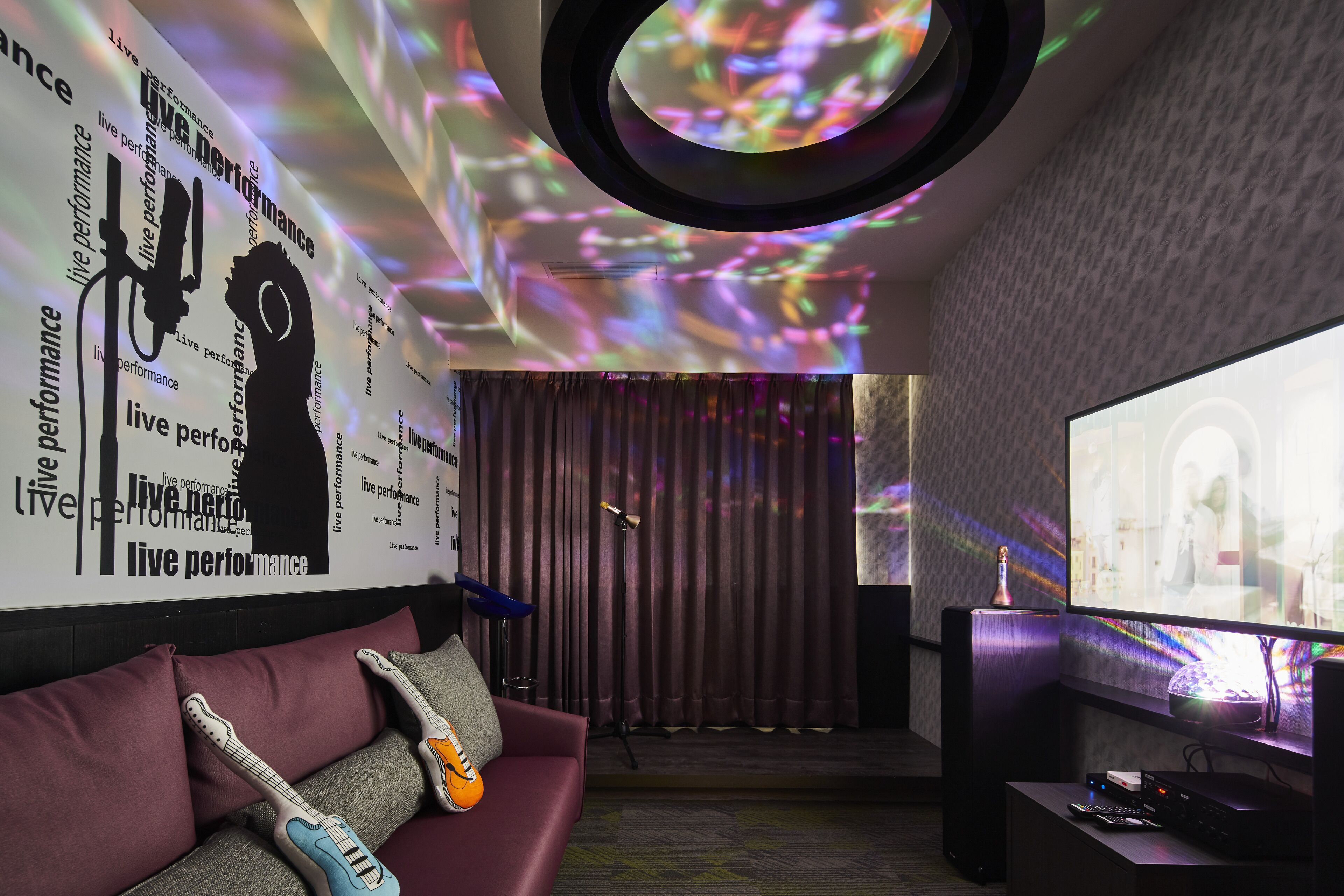 A modern living room with a large screen, a couch with a guitar, and a wall with 'live performance' text.