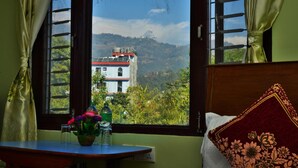 A room with a bed, a table with a vase of flowers, and a window with a view of a building and mountains. - Hotel Mandala (Pokhara)