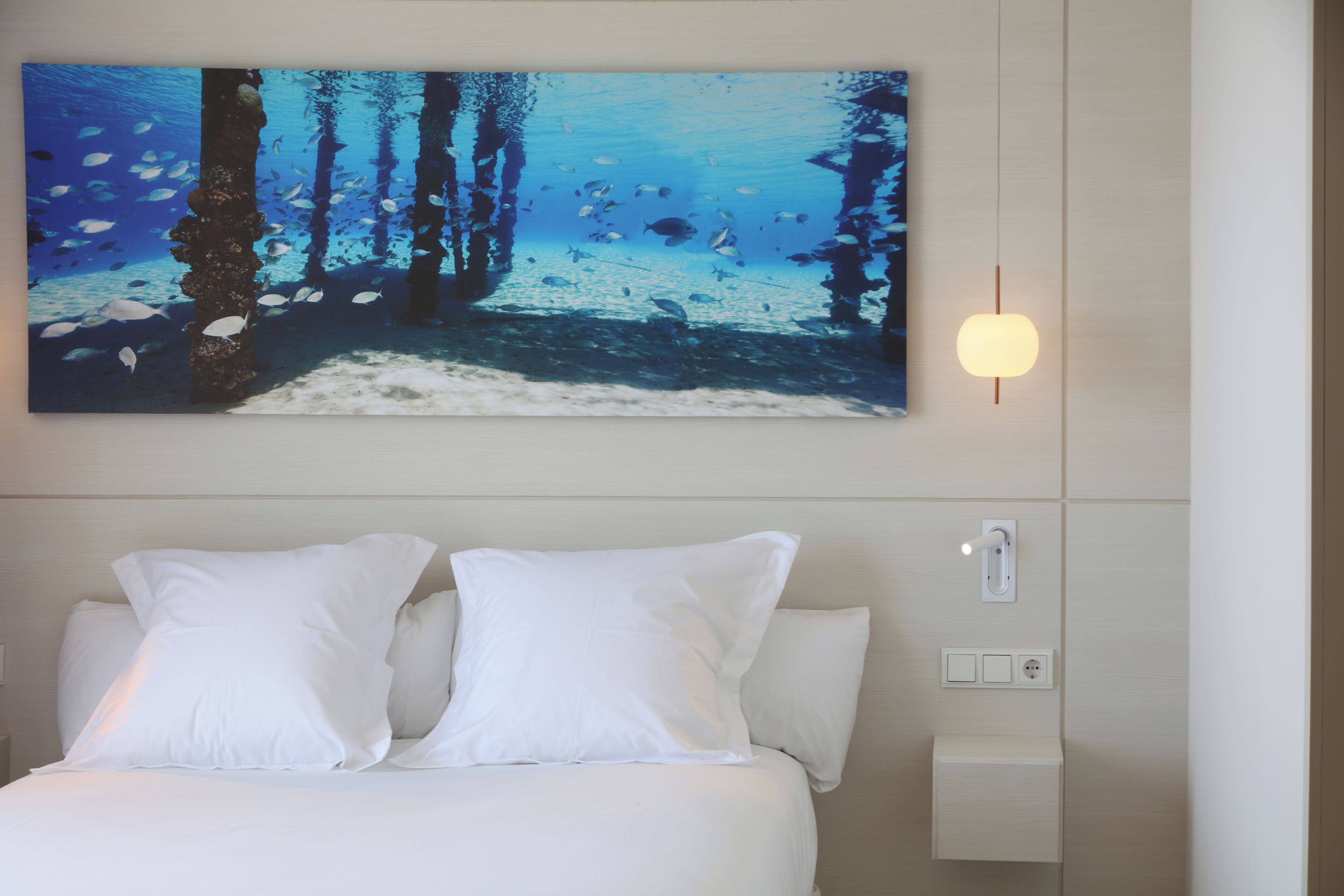 Quarto de hotel com cama, almofadas brancas, luminária fixada na parede e uma grande pintura de uma cena submarina.
