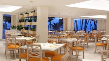 Uma sala de jantar espaçosa com várias mesas e cadeiras, grandes janelas com vista para árvores e uma parede com plantas em vasos.