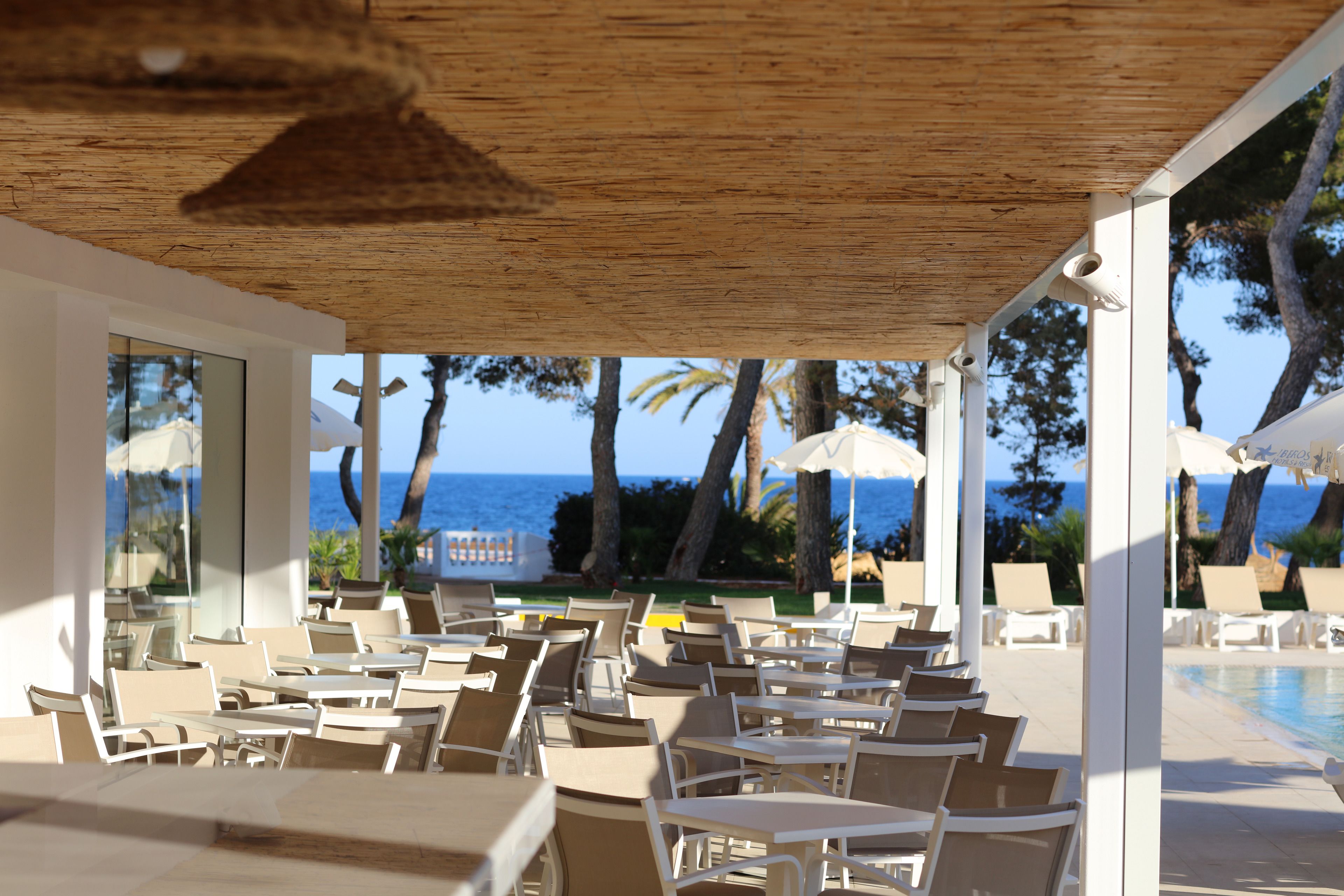 Ein überdachter Außenbereich mit Tischen und Stühlen, mit Blick auf einen Pool und das Meer.