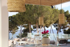Un espacio al aire libre con vista al mar, con luces colgantes de mimbre, un jarrón con flores rosadas y un candelabro de cristal.