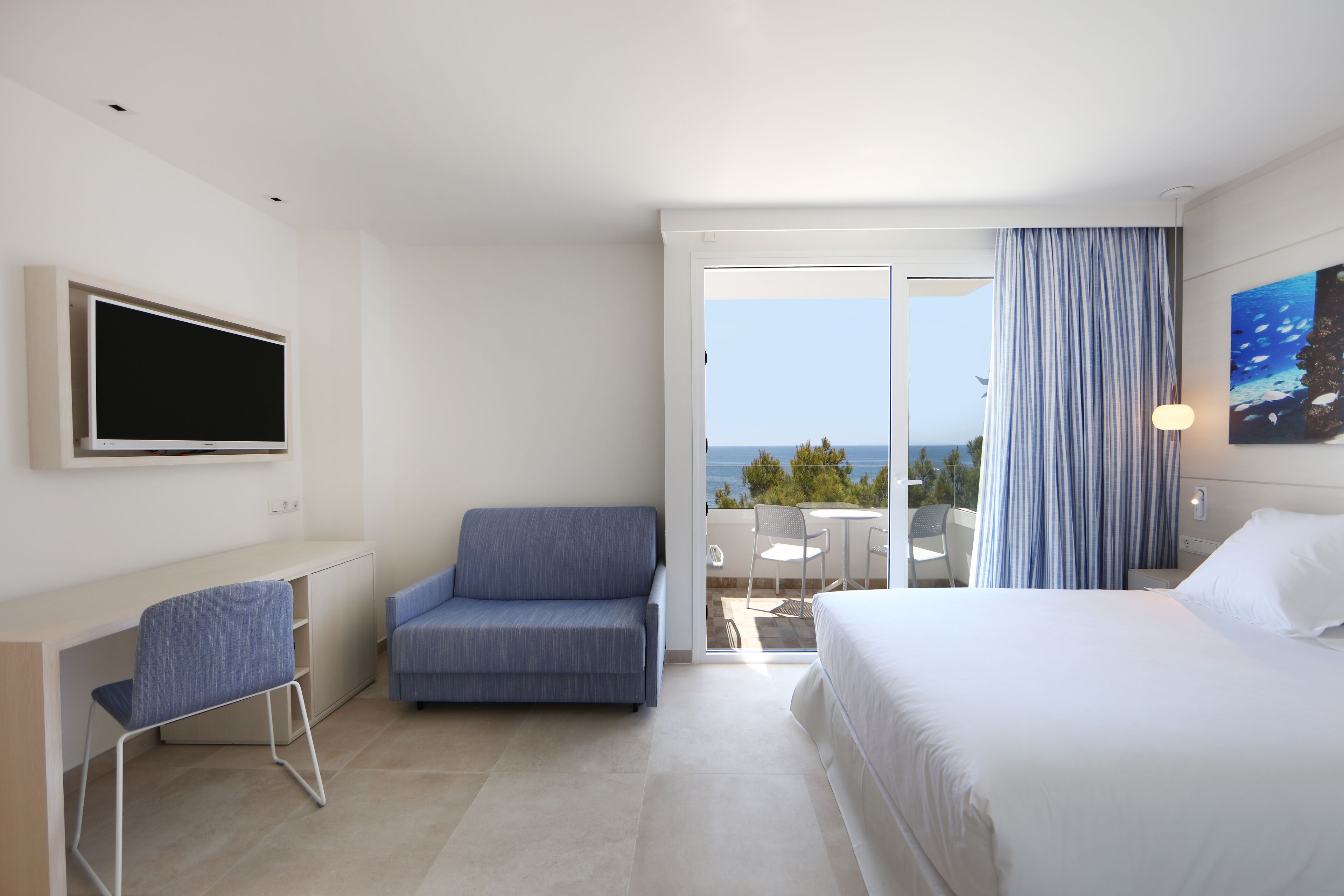 Habitación de hotel con cama, escritorio, silla, televisión y balcón con vista al mar.