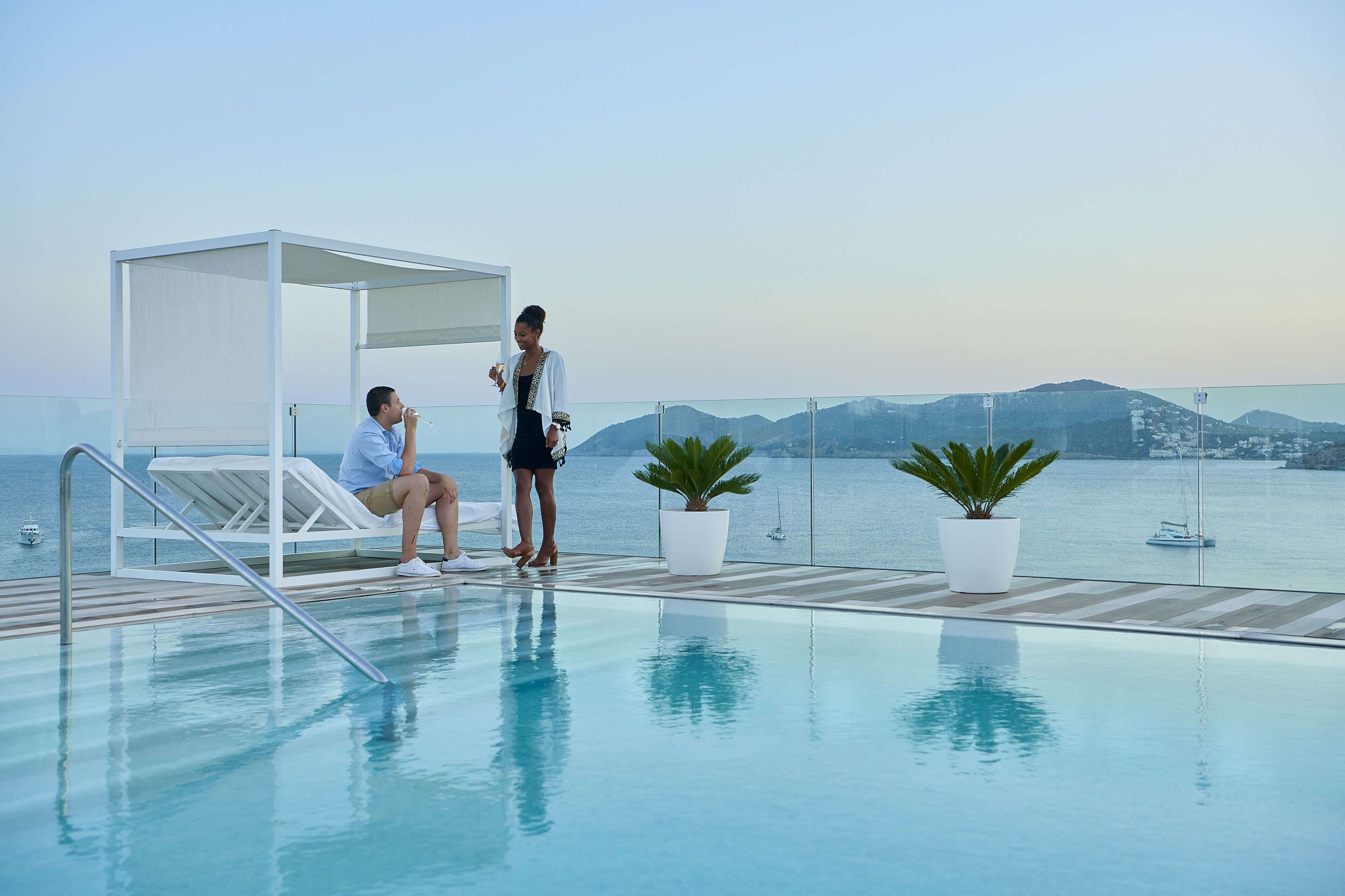 A poolside area with a glass railing, a covered seating area, and potted plants.