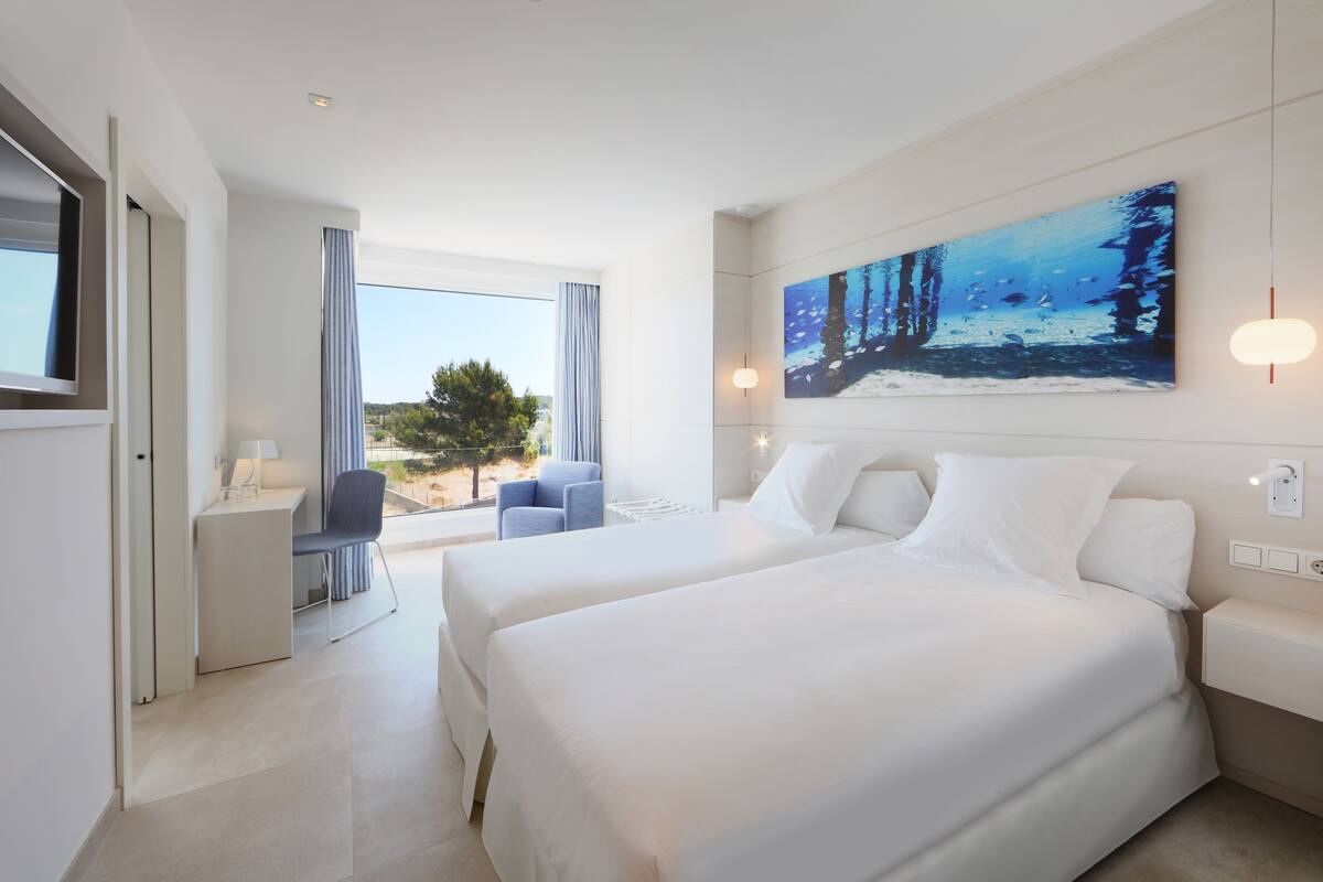 A modern hotel room with two beds, a desk, a chair, and a large window offering a view of the outdoors.