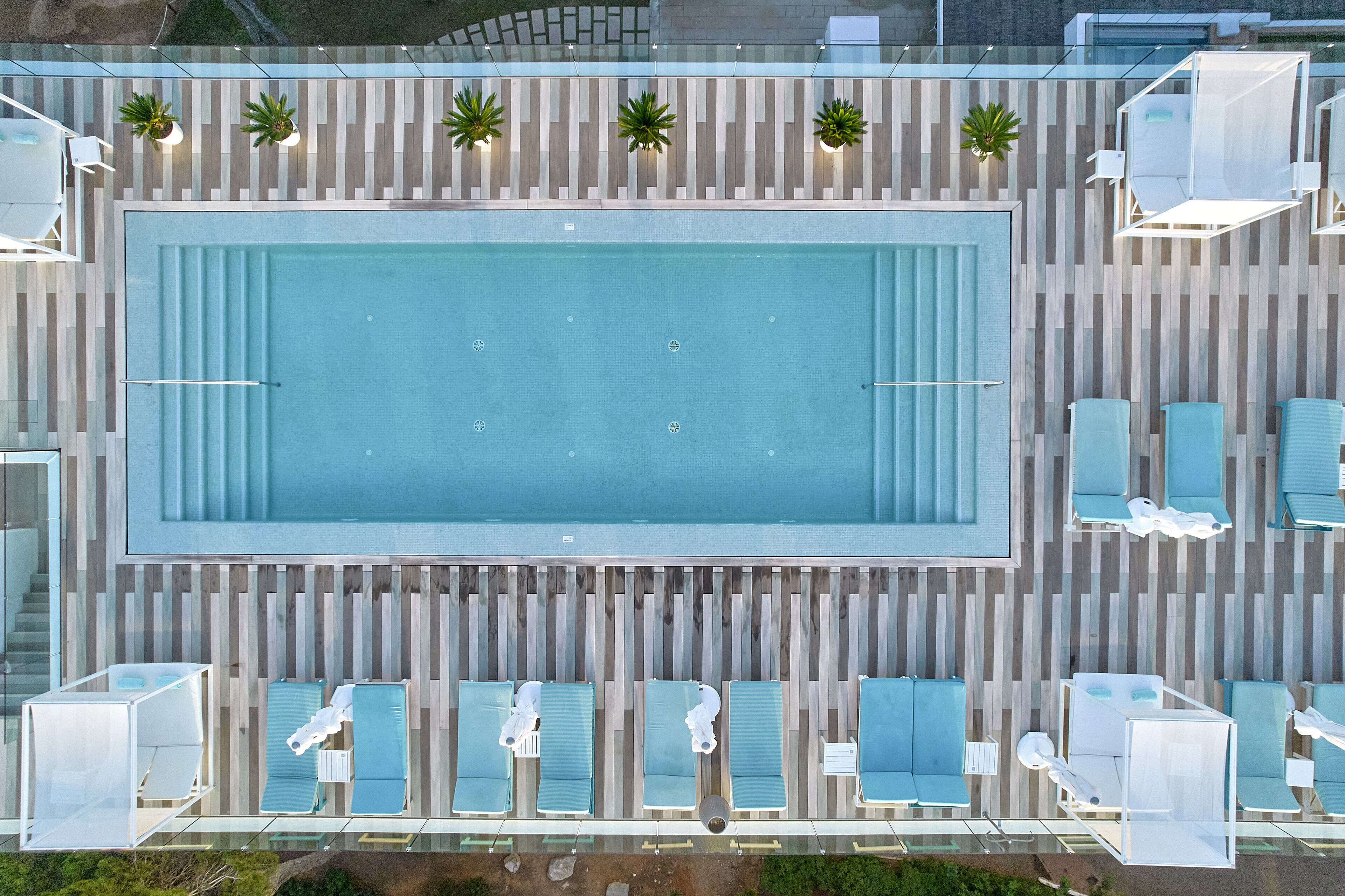 Eine Vogelperspektive auf ein rechteckiges Schwimmbecken, umgeben von Liegestühlen und Sonnenliegen, mit einer gestreiften Wand und Blumentöpfen.