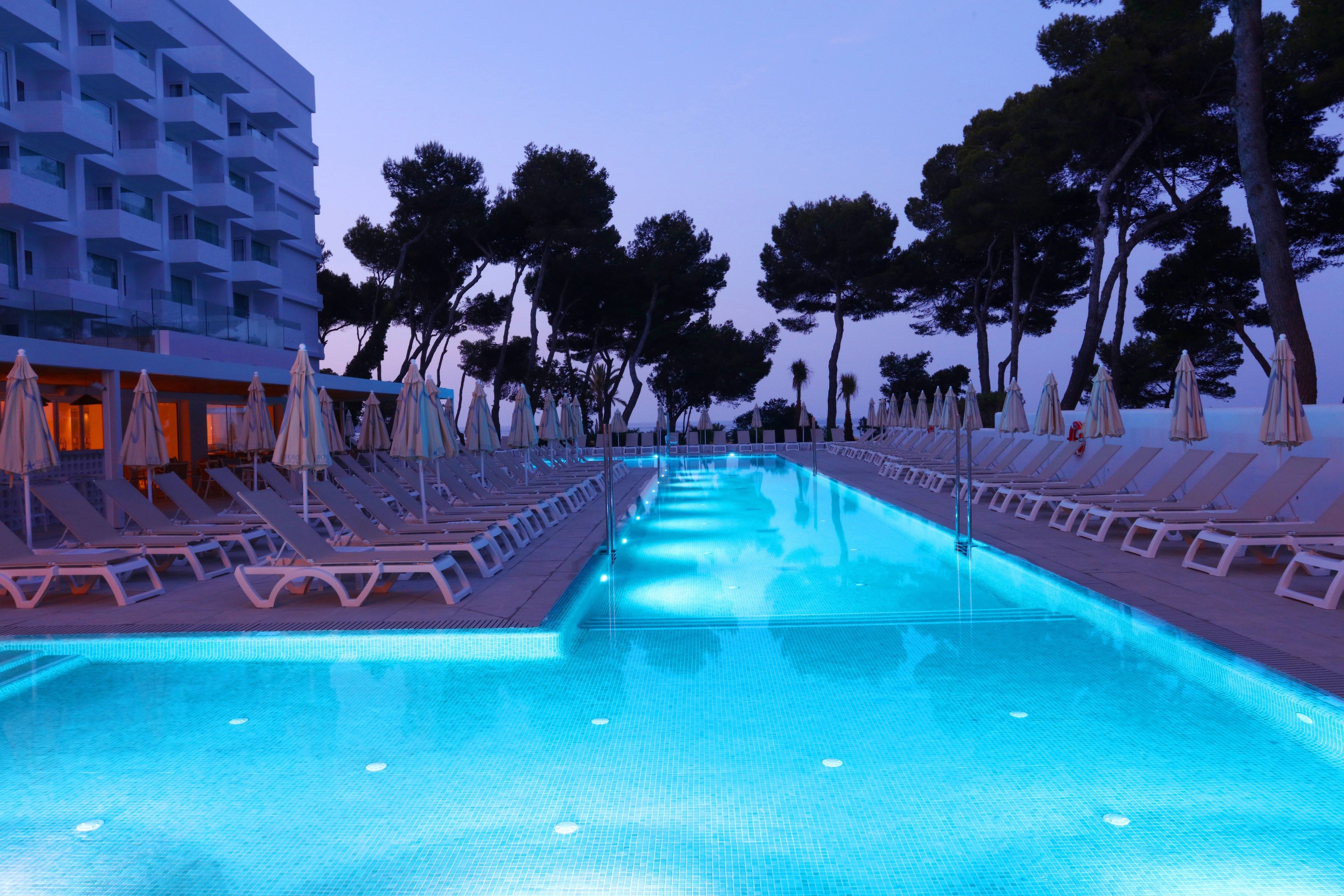 A swimming pool with lounge chairs and umbrellas, surrounded by trees and a modern building.
