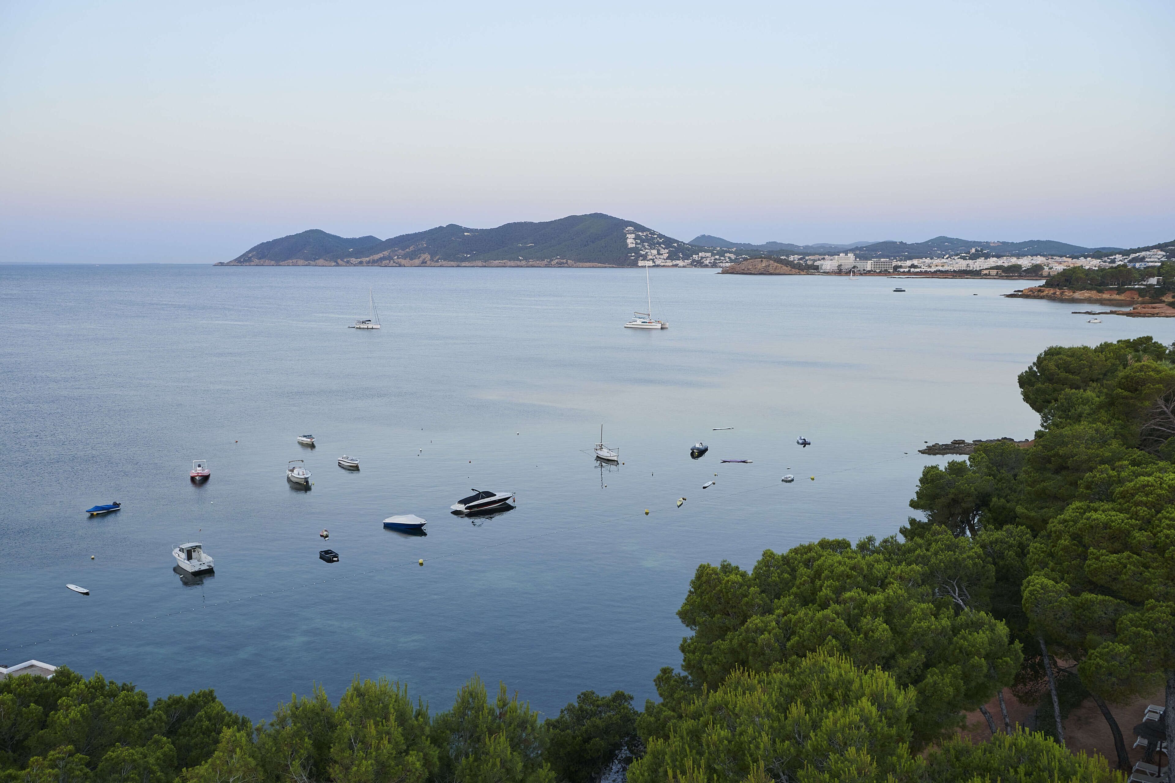 Vista costera con barcos en aguas tranquilas, una montaña distante y abundante vegetación.