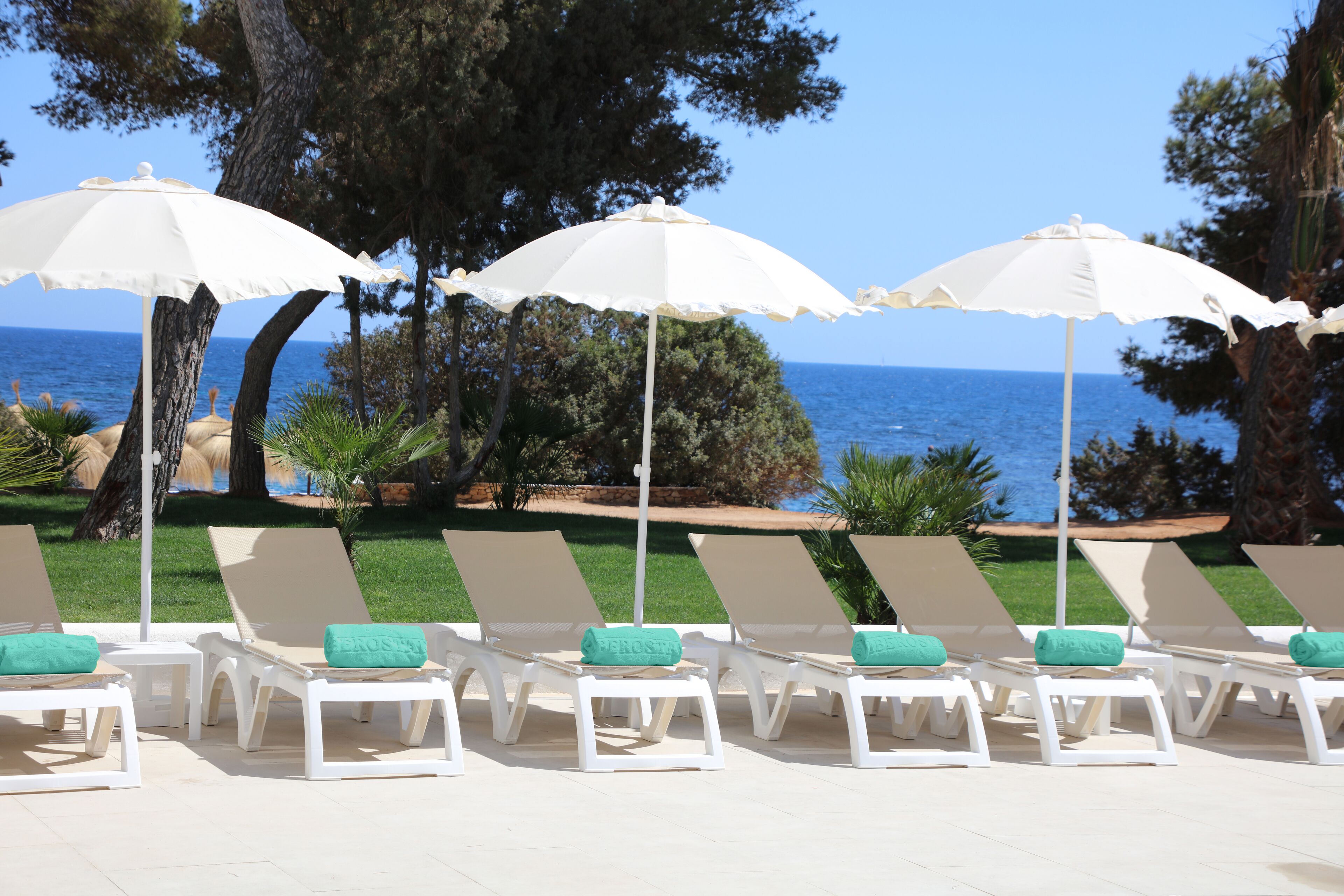 Une rangée de chaises longues blanches, dotées de coussins verts et de parasols blancs, est disposée sur une terrasse carrelée, offrant une vue sur la mer.