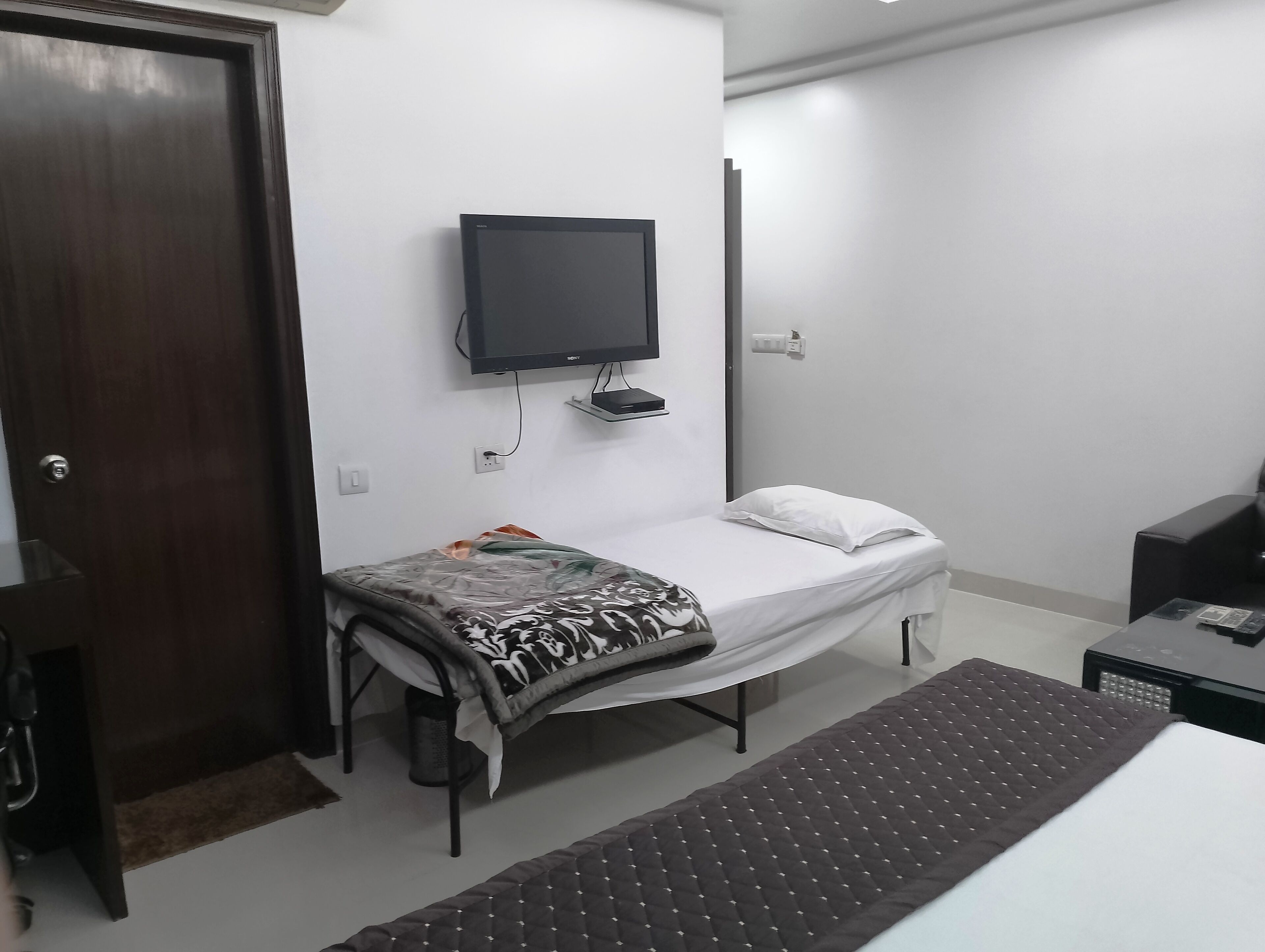 A small, clean room with a bed, a TV mounted on the wall, a desk, and a door.