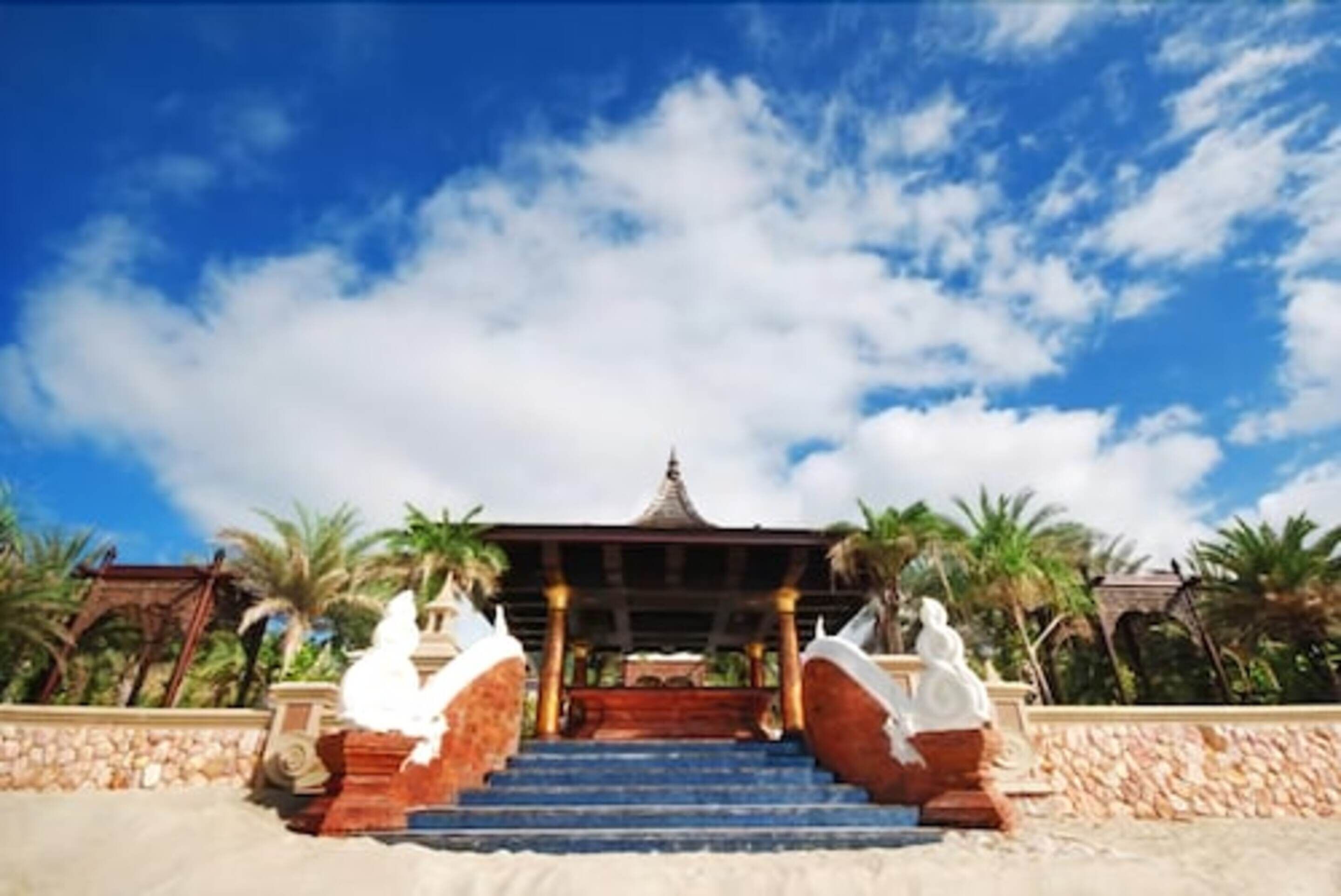 A temple with a staircase leading up to a roofed structure, surrounded by palm trees and a clear sky.