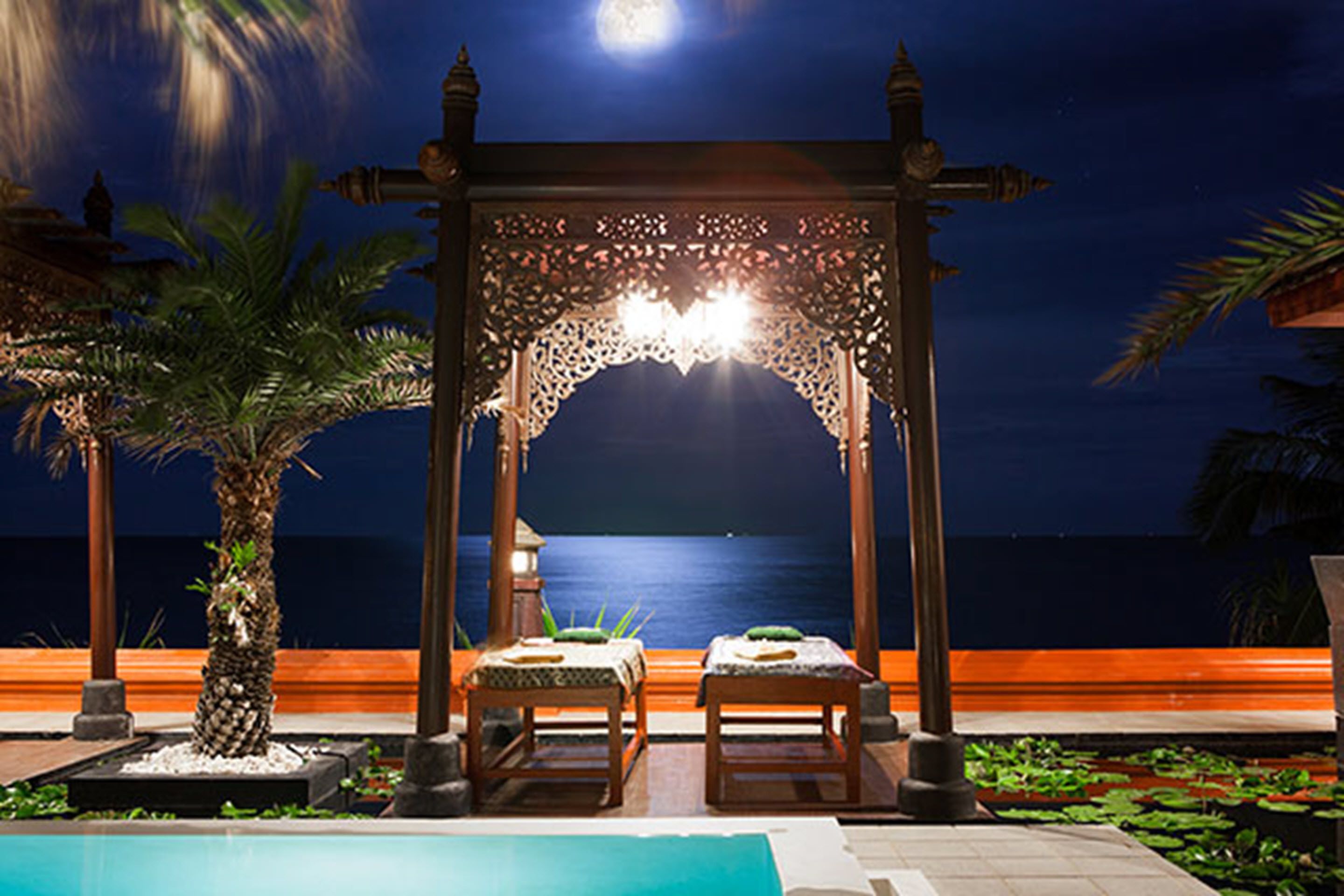A night-time view of a beachside resort with a wooden gazebo, two massage tables, and a pool.