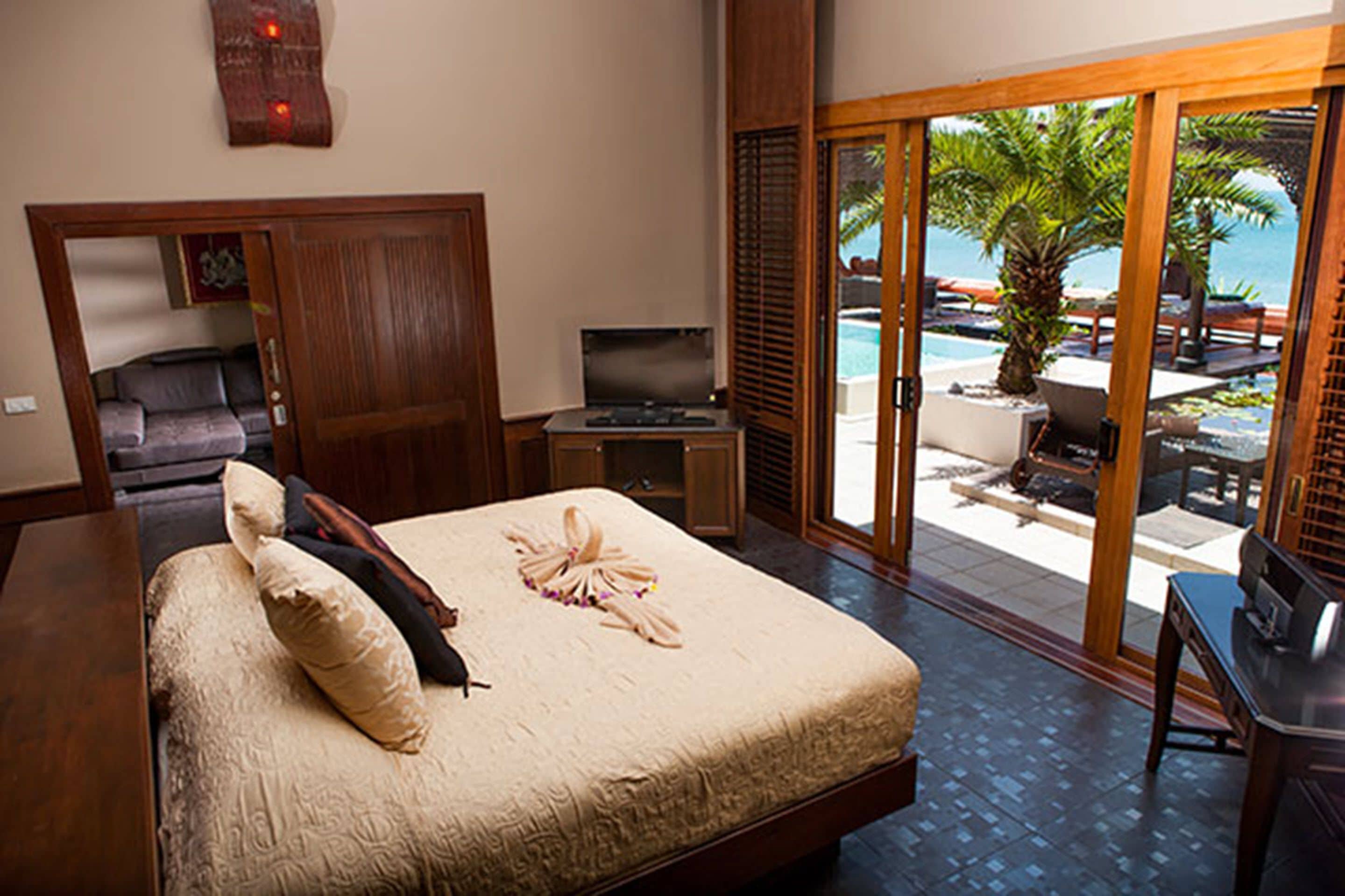A bedroom with a large bed, a television, a desk, and a view of a pool and palm trees.