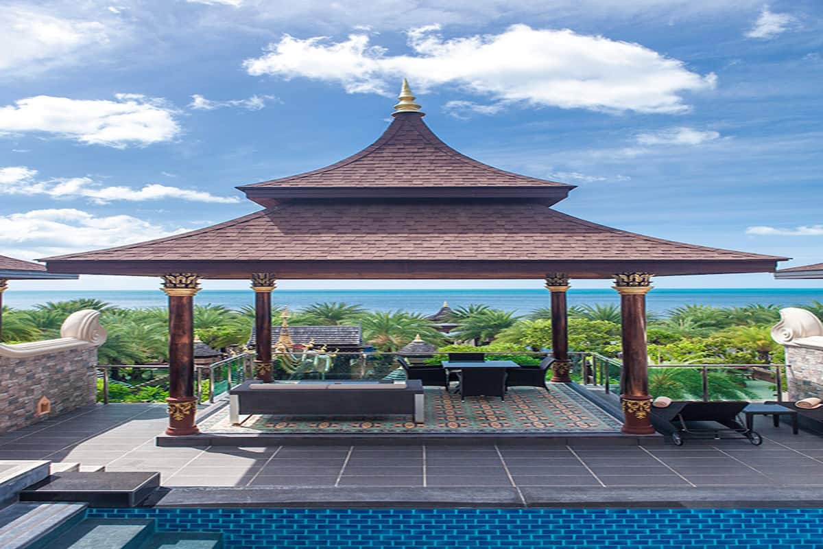 A covered outdoor seating area with a pool in the foreground, a gazebo with a pointed roof, and a view of the ocean in the background.