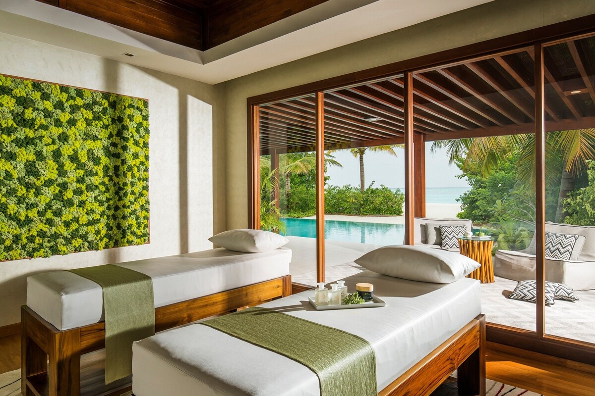 Ein Spa-Behandlungsraum mit zwei Massagetischen, einem großen Fenster mit Blick auf einen Pool und tropische Landschaft, sowie einer Holzbalkendecke.