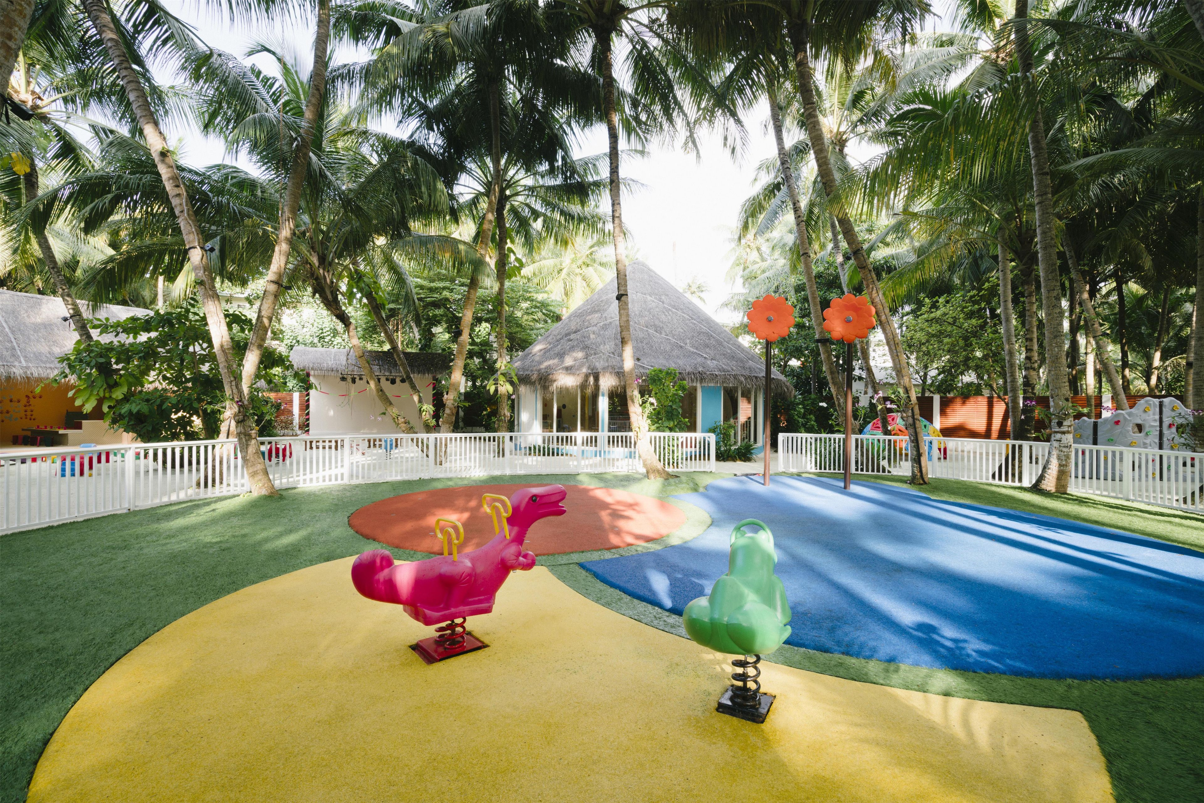 A playground with colorful play equipment, surrounded by palm trees and a white fence.