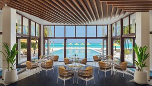 A poolside dining area with wicker chairs and tables, large windows offering a view of the ocean, and a wooden ceiling.