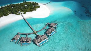Una vista aerea di bungalow sull'acqua collegati da un ponte, immersi in acque turchesi e una piccola isola ricoperta da vegetazione lussureggiante.