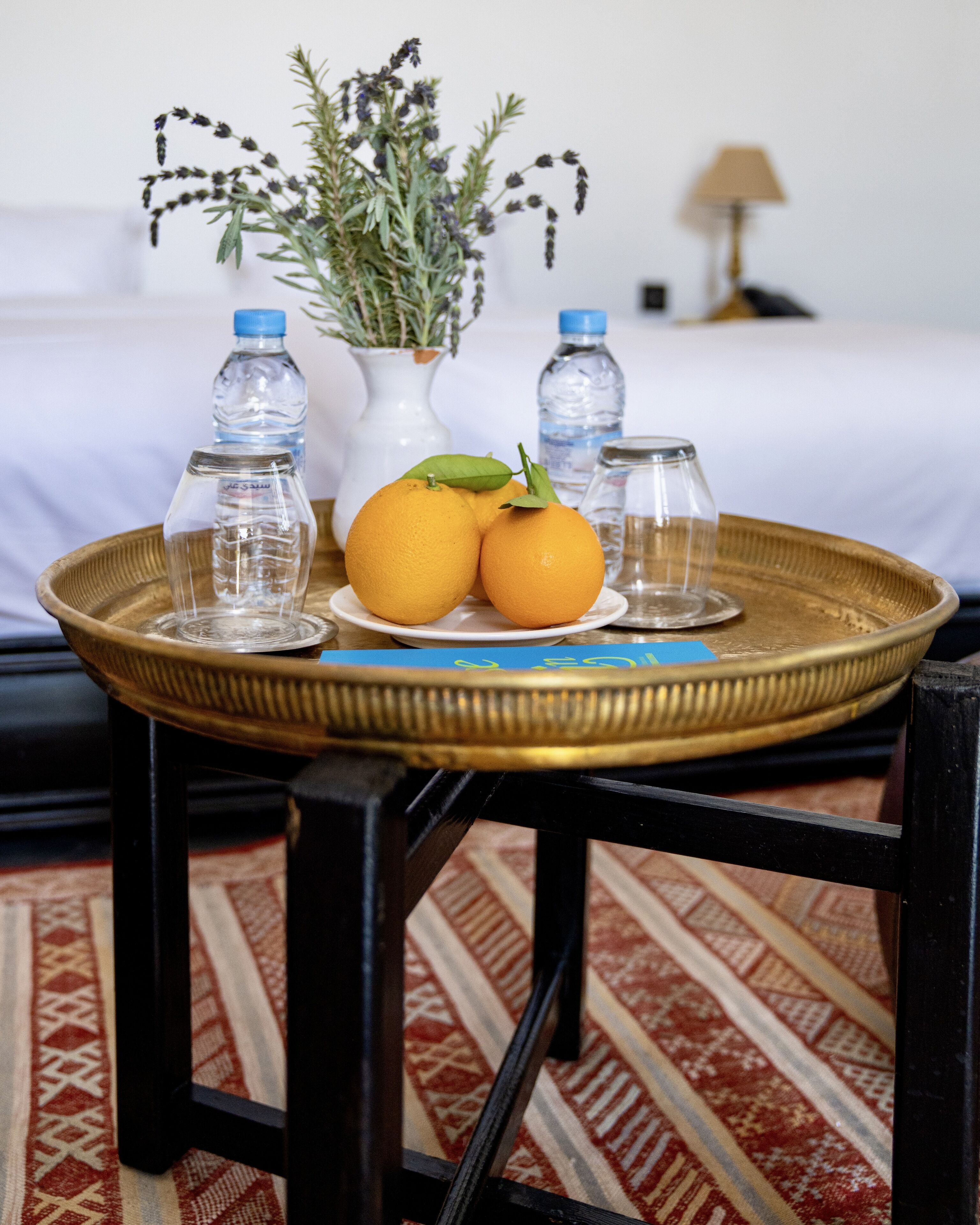Un plateau avec deux oranges et deux verres sur une table, un vase avec des fleurs et deux bouteilles d’eau en arrière-plan.