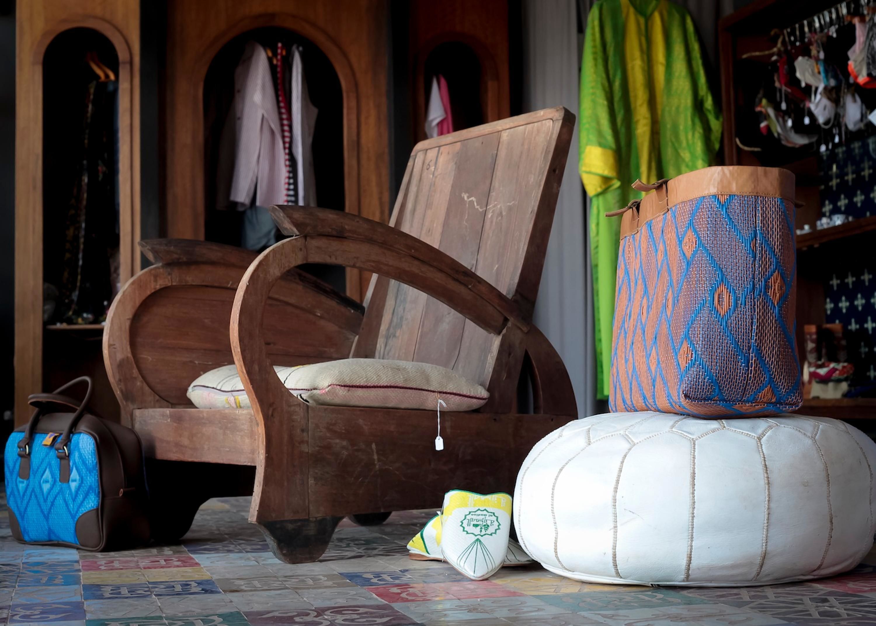 Una stanza con una sedia di legno, una borsa blu, un pouf bianco e un contenitore decorato.
