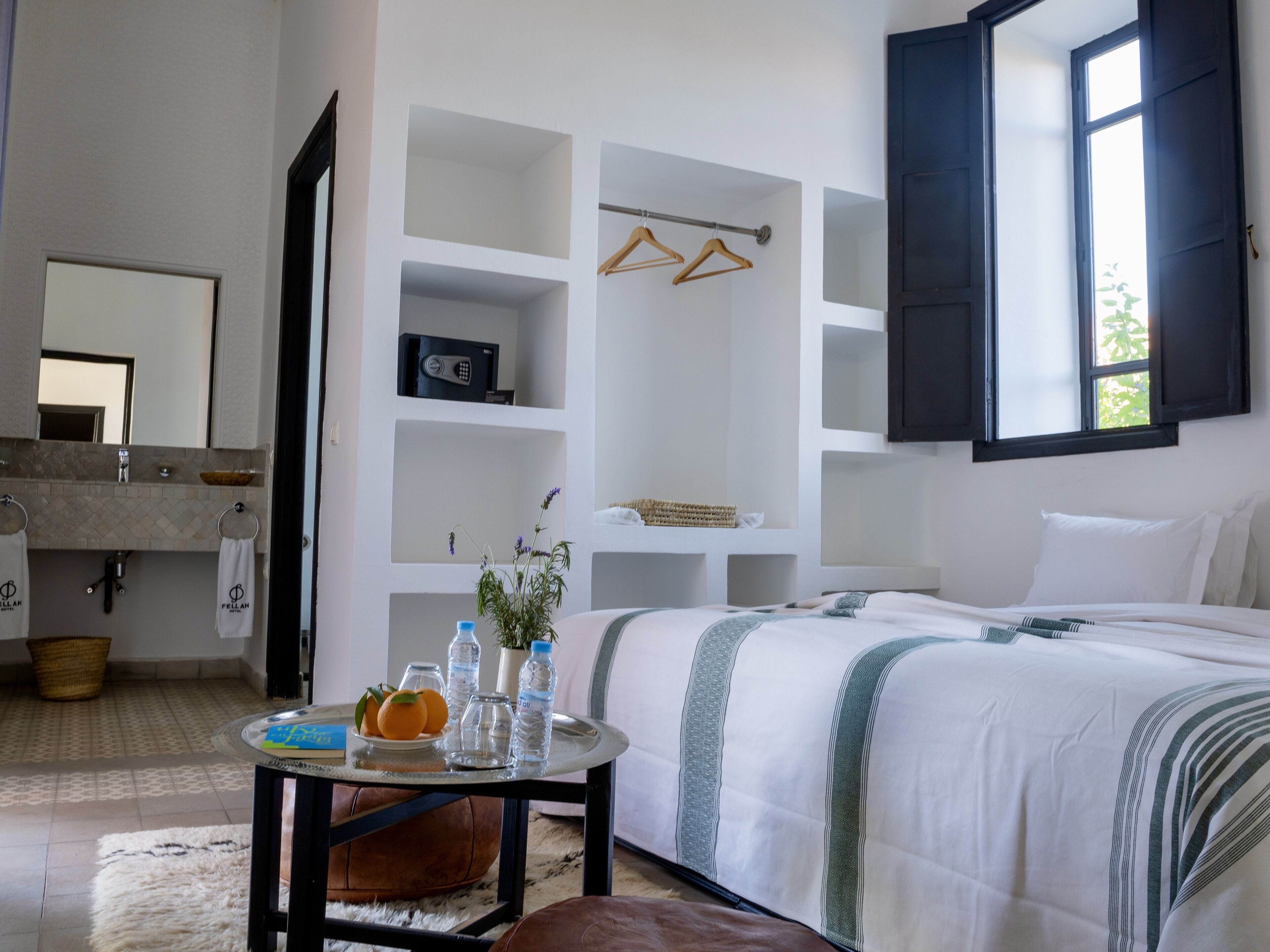 A hotel room with a bed, a round table with a vase and oranges, a mirror, a sink, and a window with shutters.