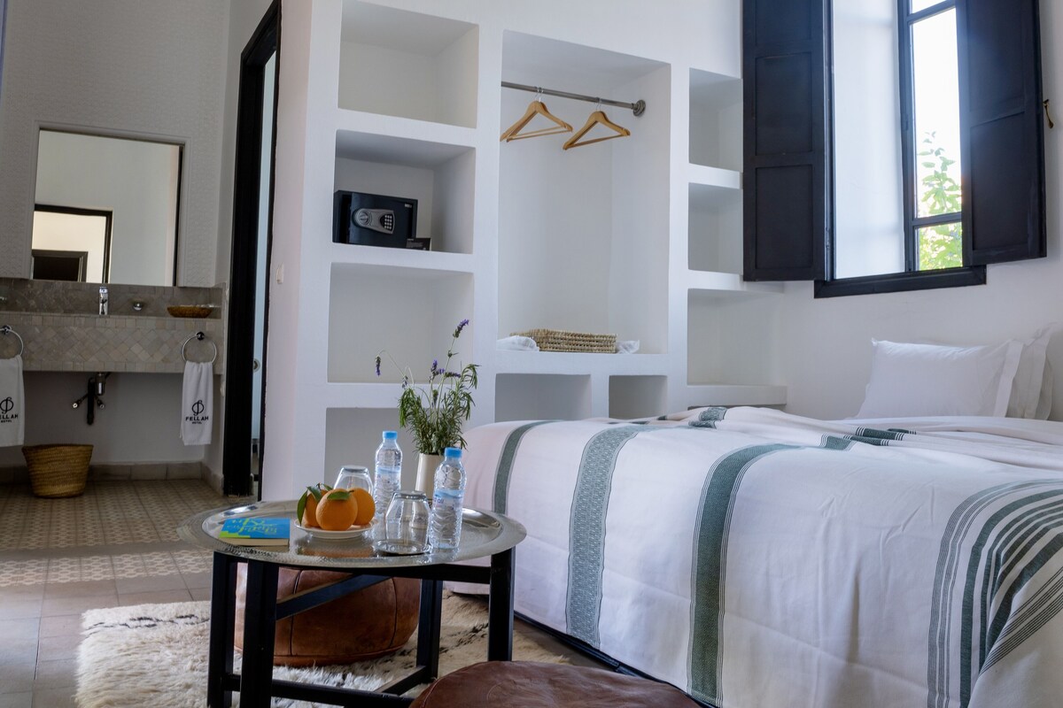 A hotel room with a bed, a round table with a vase and oranges, a mirror, a sink, and a window with shutters.