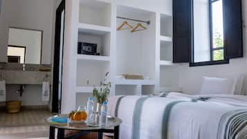 A hotel room with a bed, a round table with a vase and oranges, a mirror, a sink, and a window with shutters.