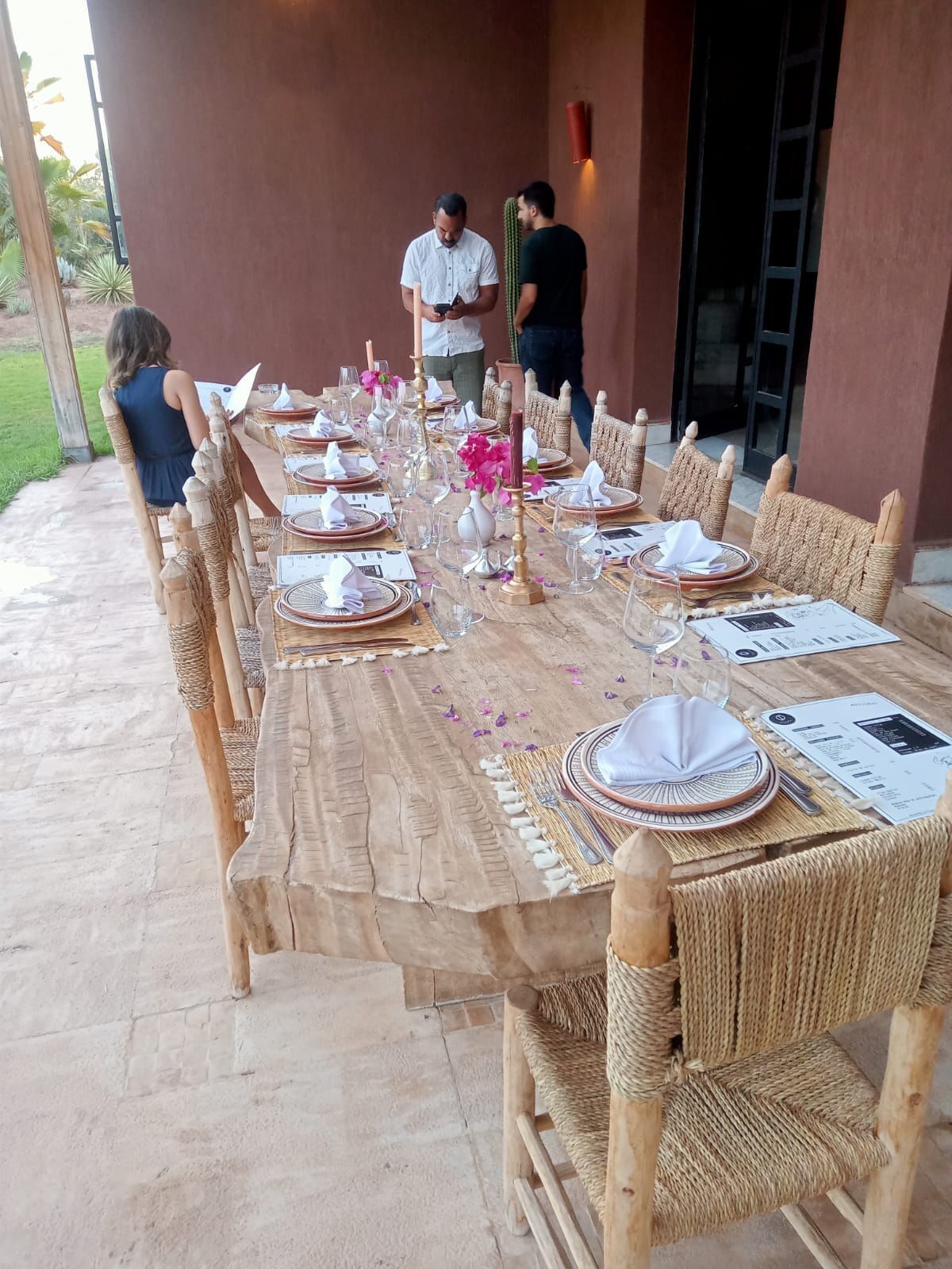 Une longue table à manger installée à l’extérieur, avec des chaises en osier, un menu et un centre de table composé d’une bougie.