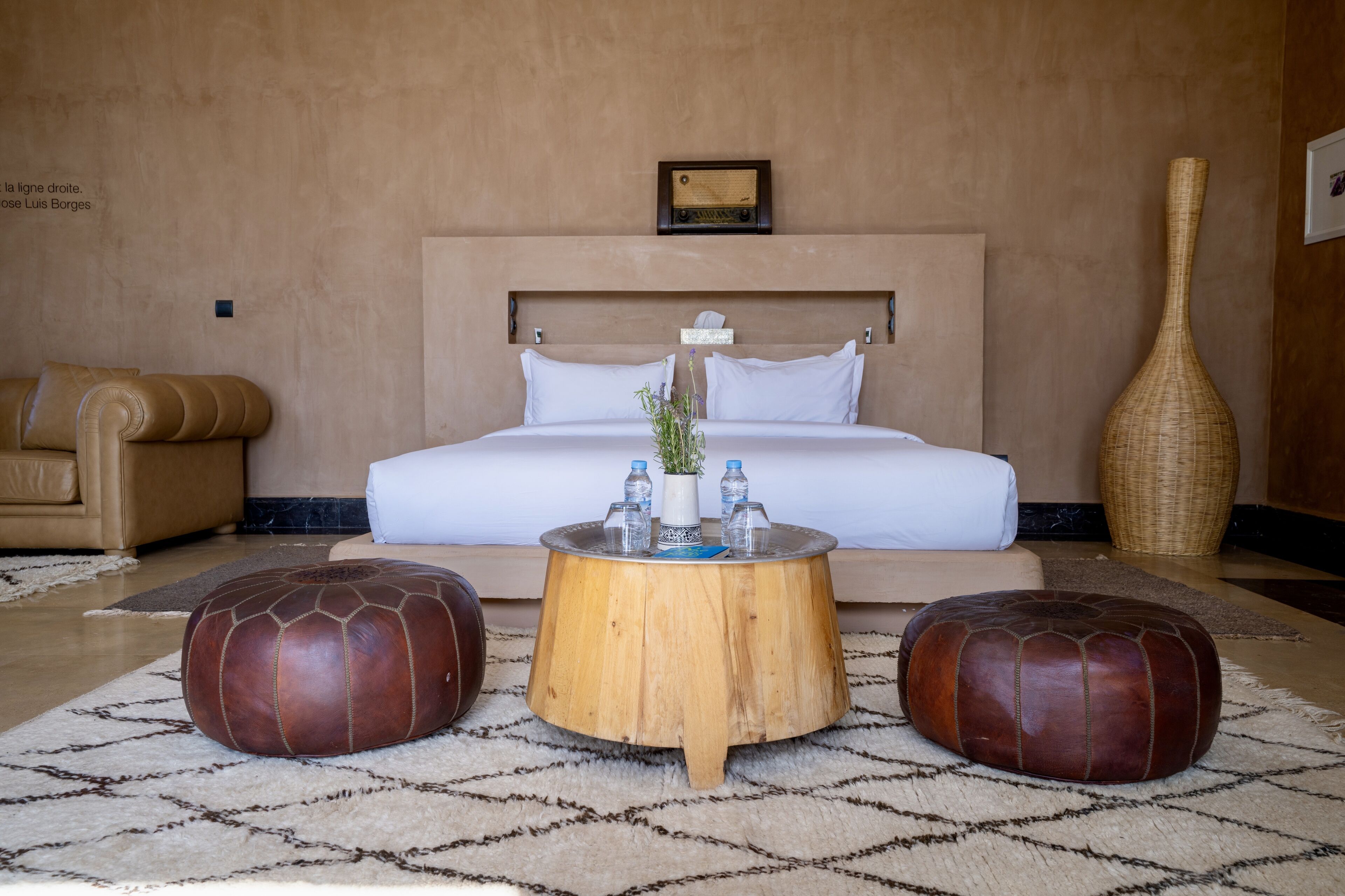 A bedroom with a bed, a wooden coffee table, two leather ottomans, a vase with plants, and a tall vase on the side.