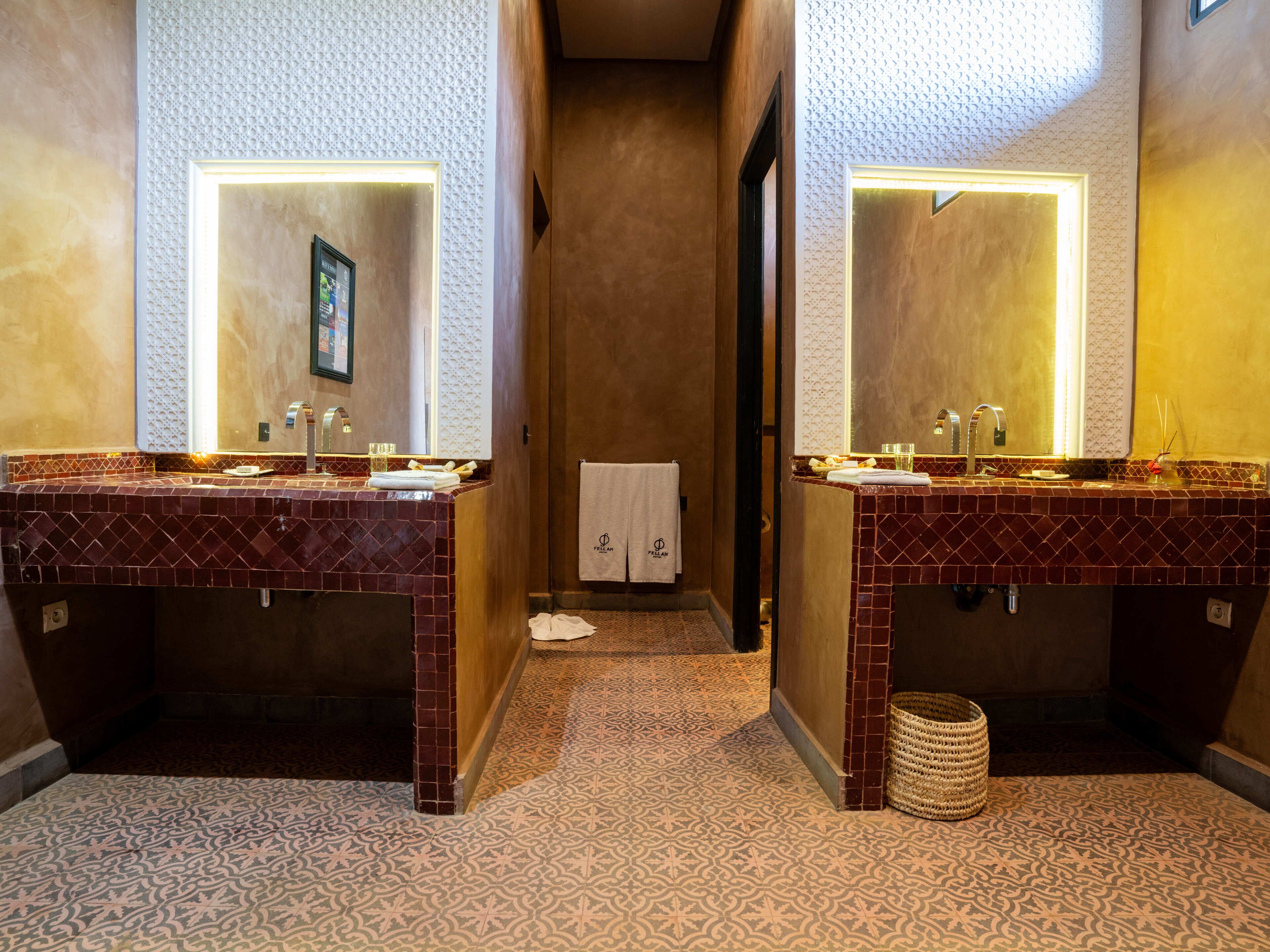 A hotel bathroom with two sinks, each with a mirror and towel. The floor has patterned tiles, and there is a trash bin near the sinks.