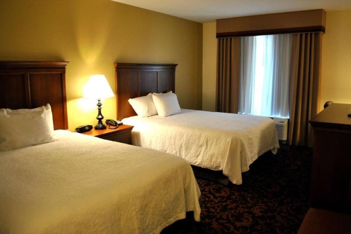 A hotel room with two beds, a bedside table with a lamp, a telephone, and a window with curtains.