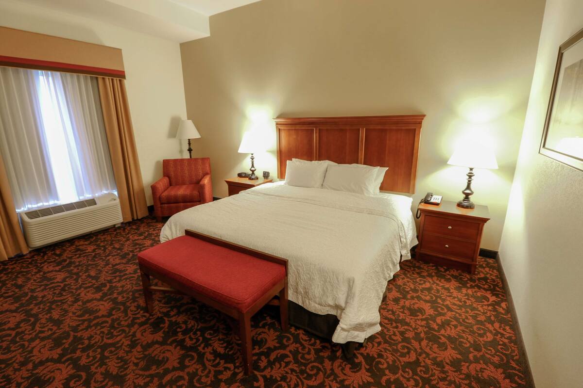 A hotel room with a large bed, two bedside tables with lamps, a red chair, a bench, and a window with curtains.