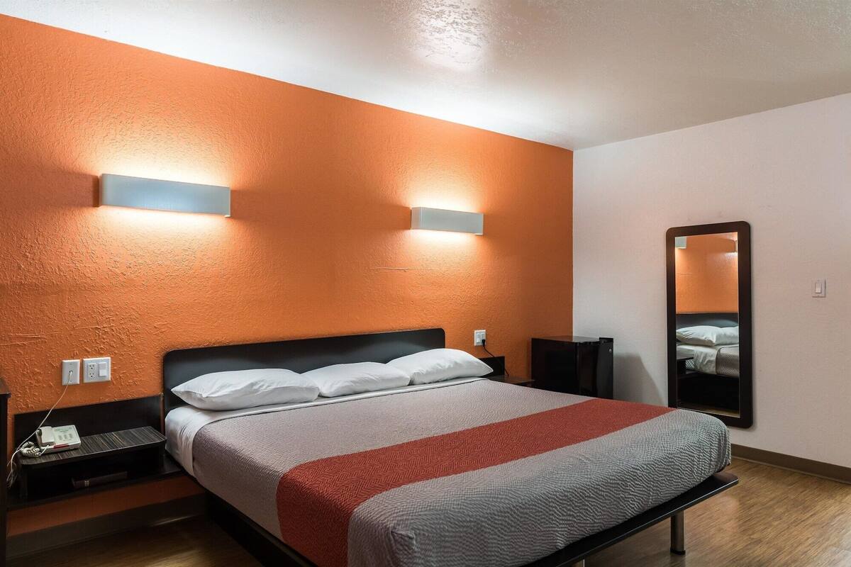 A hotel room with a bed, a nightstand, a bedside table, a mirror, and a wall-mounted light fixture.