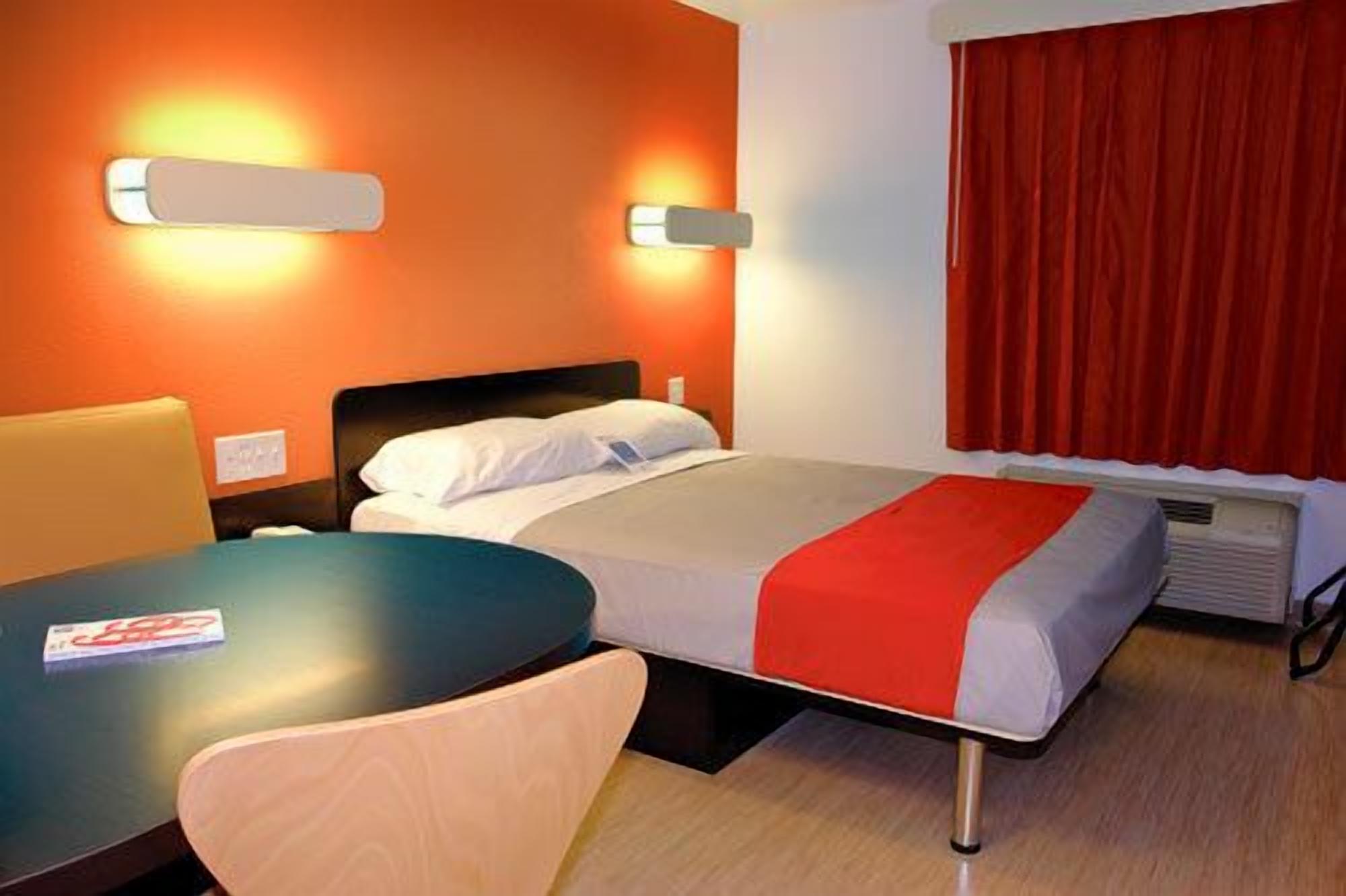 A hotel room with a bed, a table, and a chair. There are two wall-mounted lights and a window with red curtains.