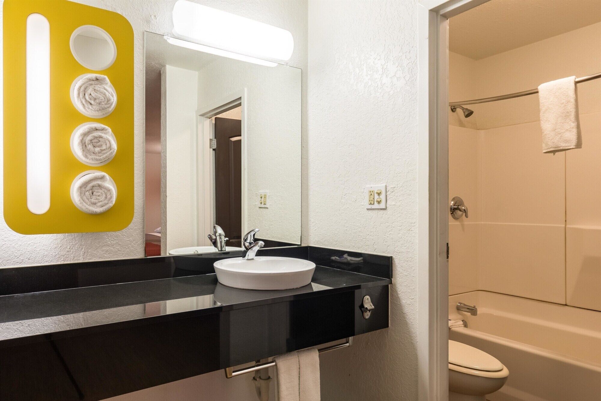 Un baño con encimera negra, lavamanos blanco y un espejo. Hay una zona de ducha con una toalla colgada en la puerta.