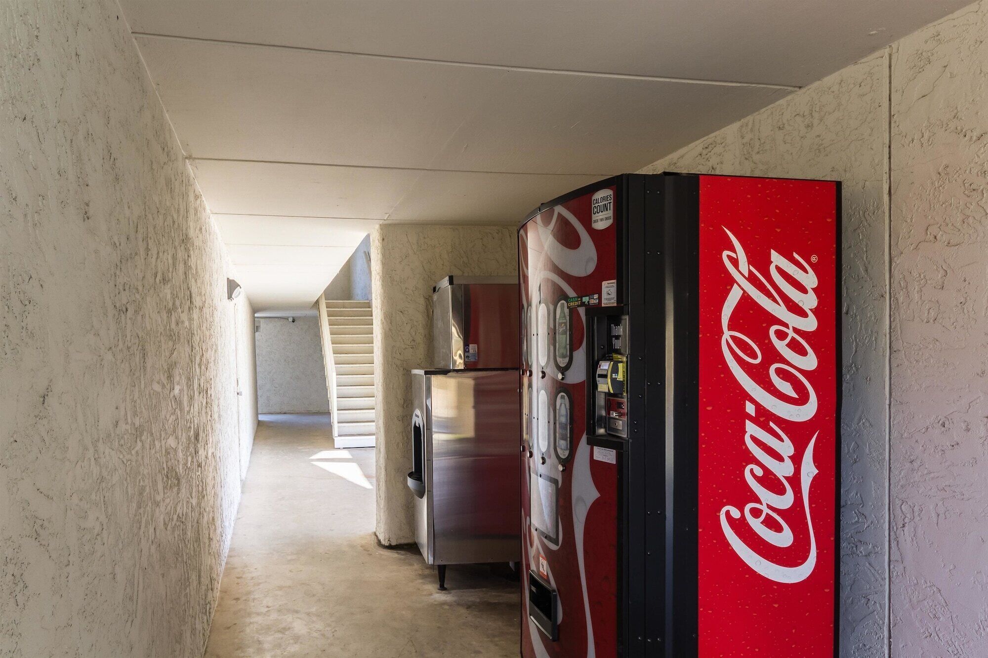 Un pasillo con una máquina expendedora de Coca-Cola, un refrigerador y una escalera que sube a otro piso.
