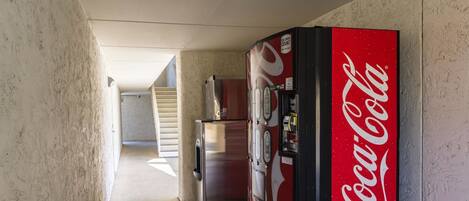 Un pasillo con una máquina expendedora de Coca-Cola, un refrigerador y una escalera que sube a otro piso.