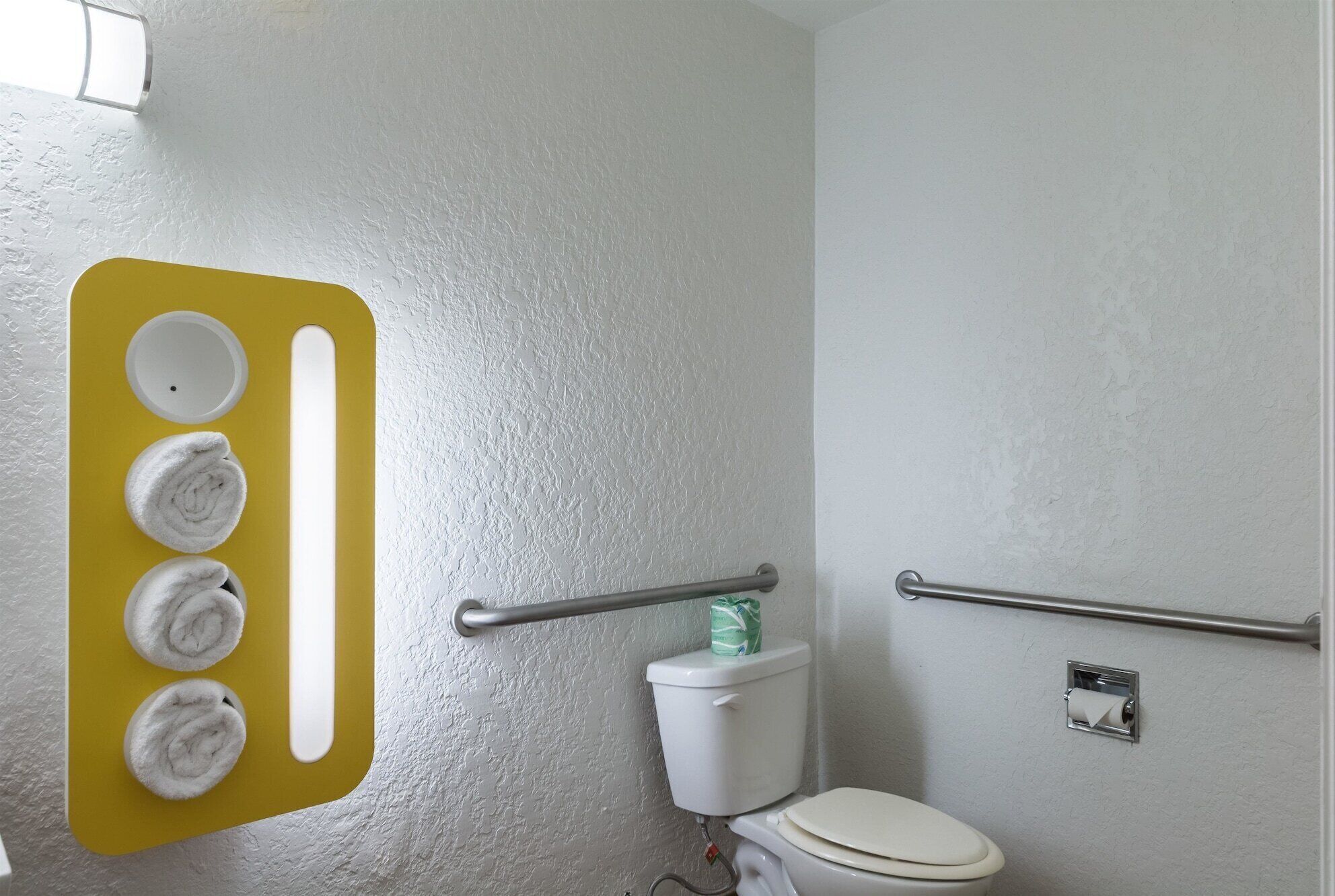 Een badkamer met een wit toilet, een geel handdoekrek met vier handdoeken, een handgreep en een toiletpapierhouder.