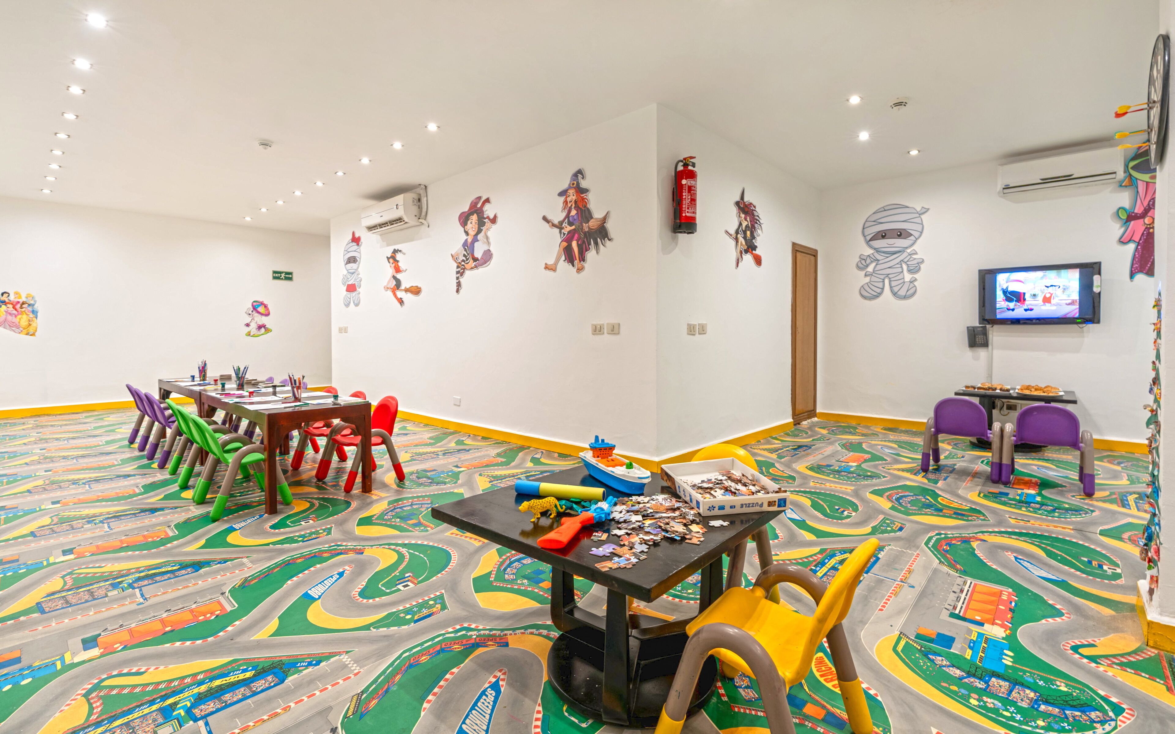 A children's playroom with colorful floor designs, a table with puzzles, and a television on the wall.