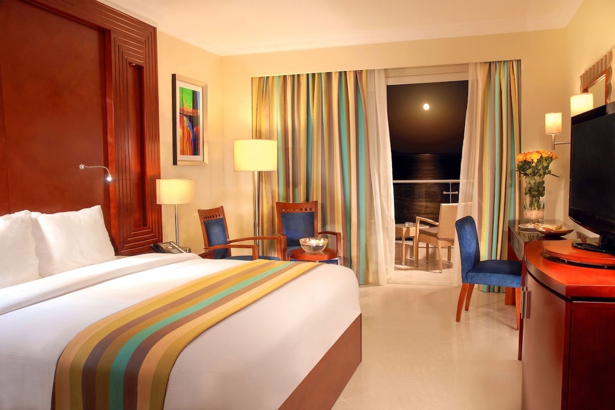 A hotel room with a large bed, a desk with a television, a chair, a small table, and a view of the outside through a window with curtains.