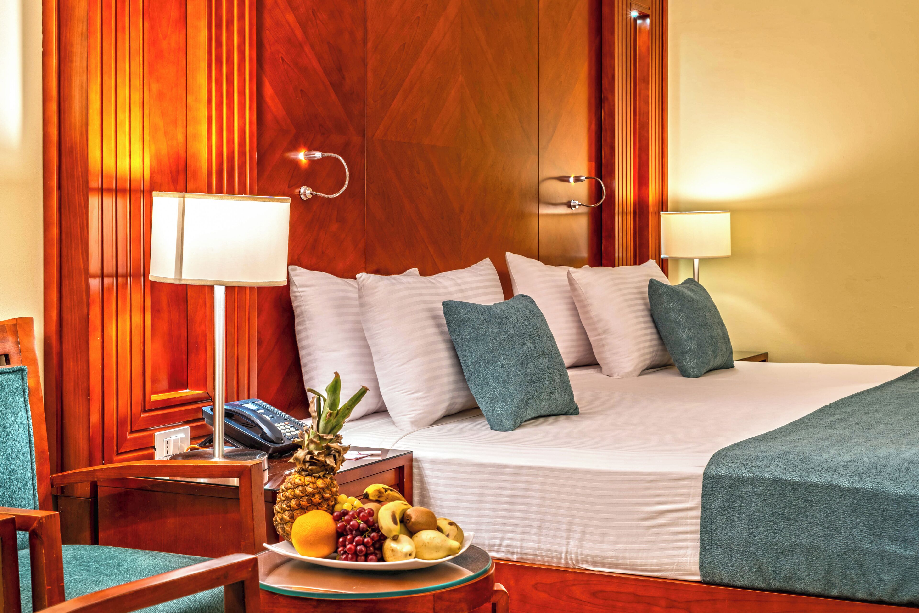 A hotel room with a bed, bedside lamps, a wooden headboard, a small table with fruit, and a chair.