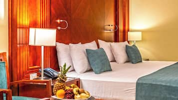 A hotel room with a bed, bedside lamps, a wooden headboard, a small table with fruit, and a chair.