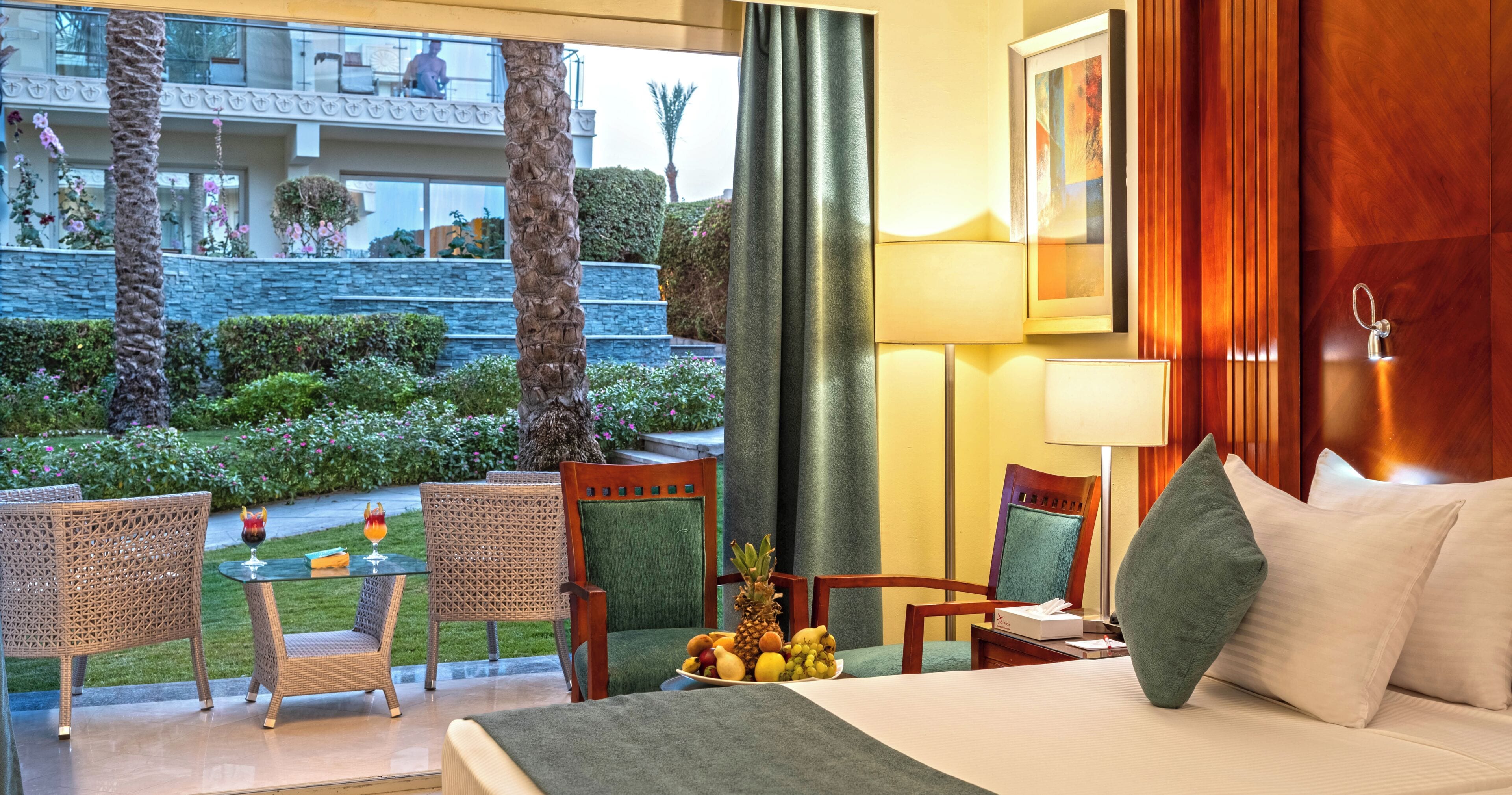 A hotel room with a bed, a table with fruit, two wicker chairs, a view of a garden, and a balcony.