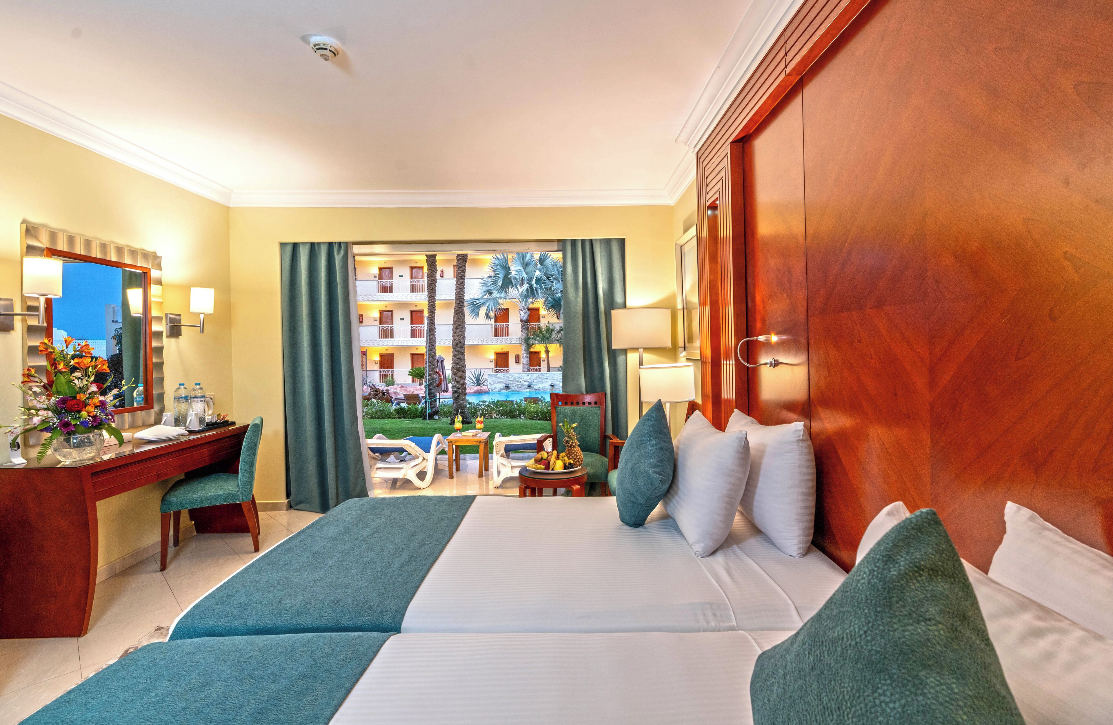 A hotel room with a large bed, a desk, a chair, a small table with fruit, and a view of the hotel building and palm trees.