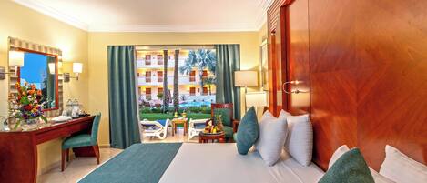 A hotel room with a large bed, a desk, a chair, a small table with fruit, and a view of the hotel building and palm trees.